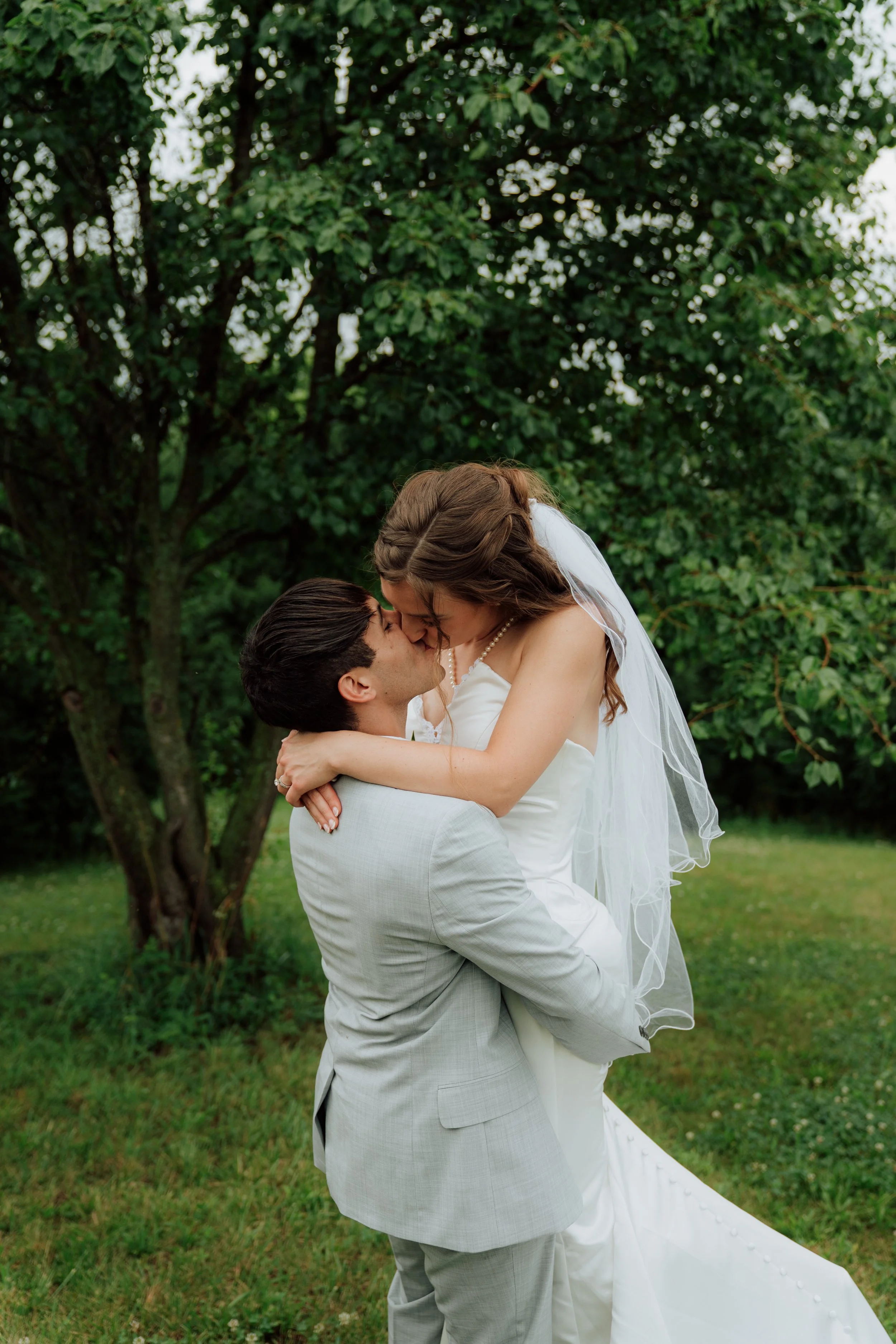 A bride and groom share a kiss outdoors, with the groom lifting the bride, both dressed in wedding attire, surrounded by green trees and grass.
