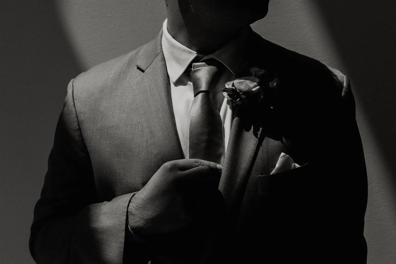 A man dressed in a suit with a boutonniere, holding a rose up to his chest in a black and white photo.
