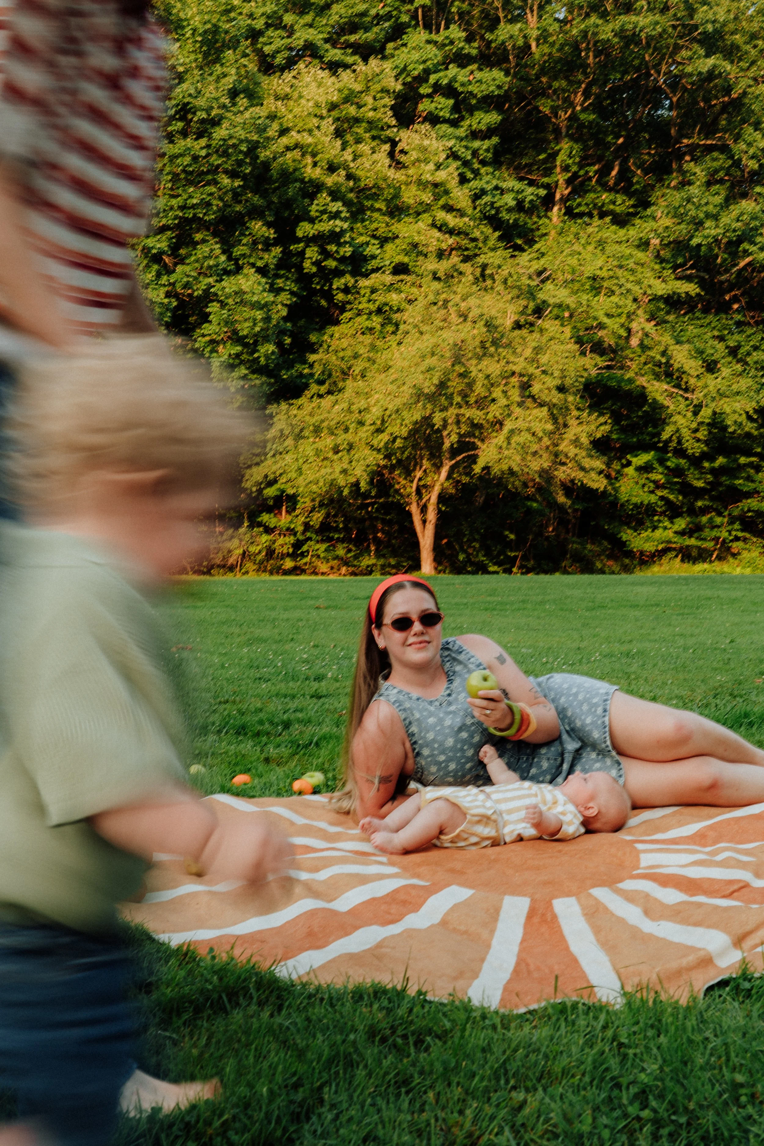 A woman with sunglasses lays on a blanket on the grass, holding a green apple. A baby lies beside her, and a man in the foreground is partially blurred. The background features large trees and a lush green lawn.