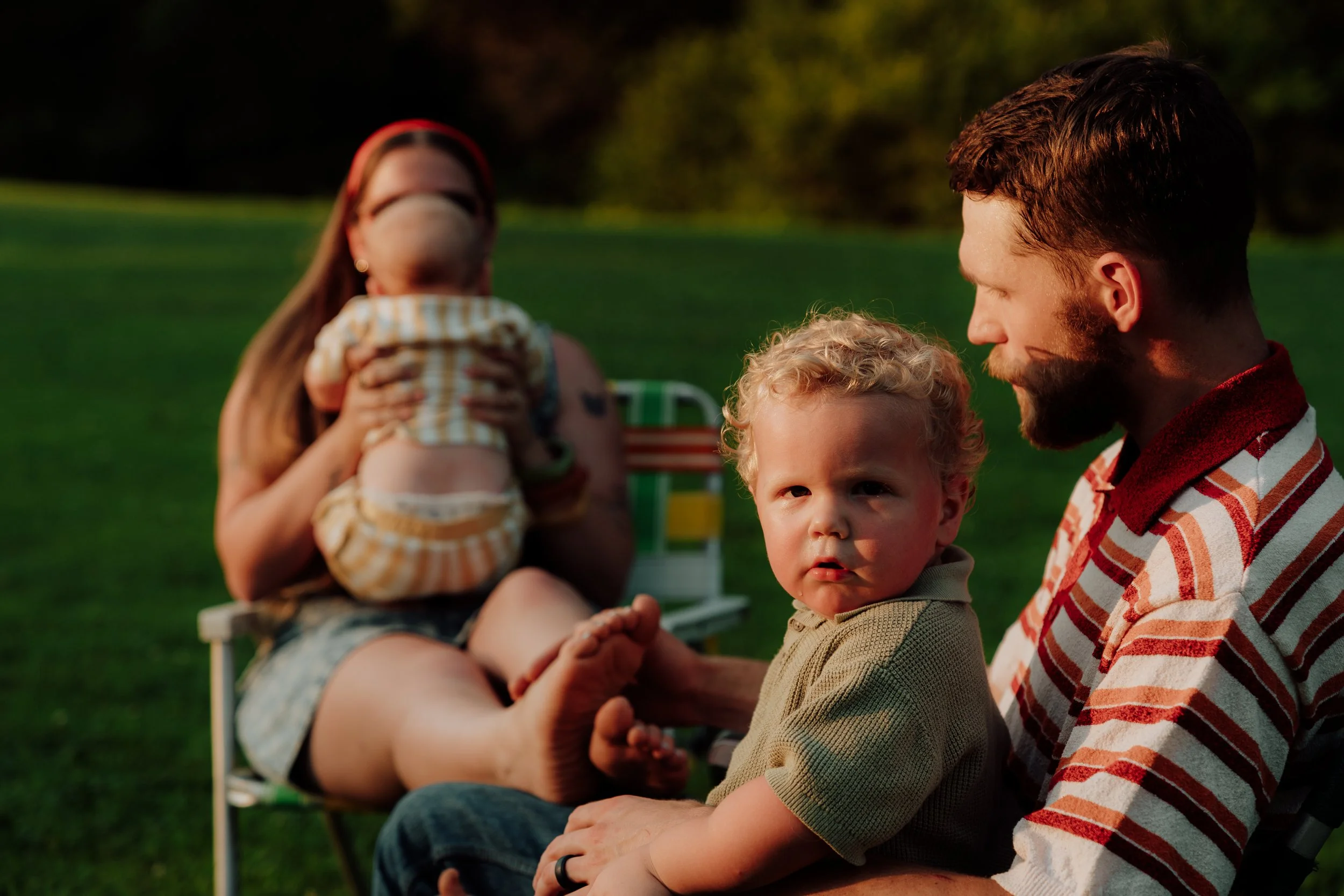 A man with a beard in a red and white striped shirt holding a toddler with curly blonde hair, sitting outdoors on grass. A woman in the background is holding a baby with a red headband and a checkered outfit. The scene is taken during sunset.