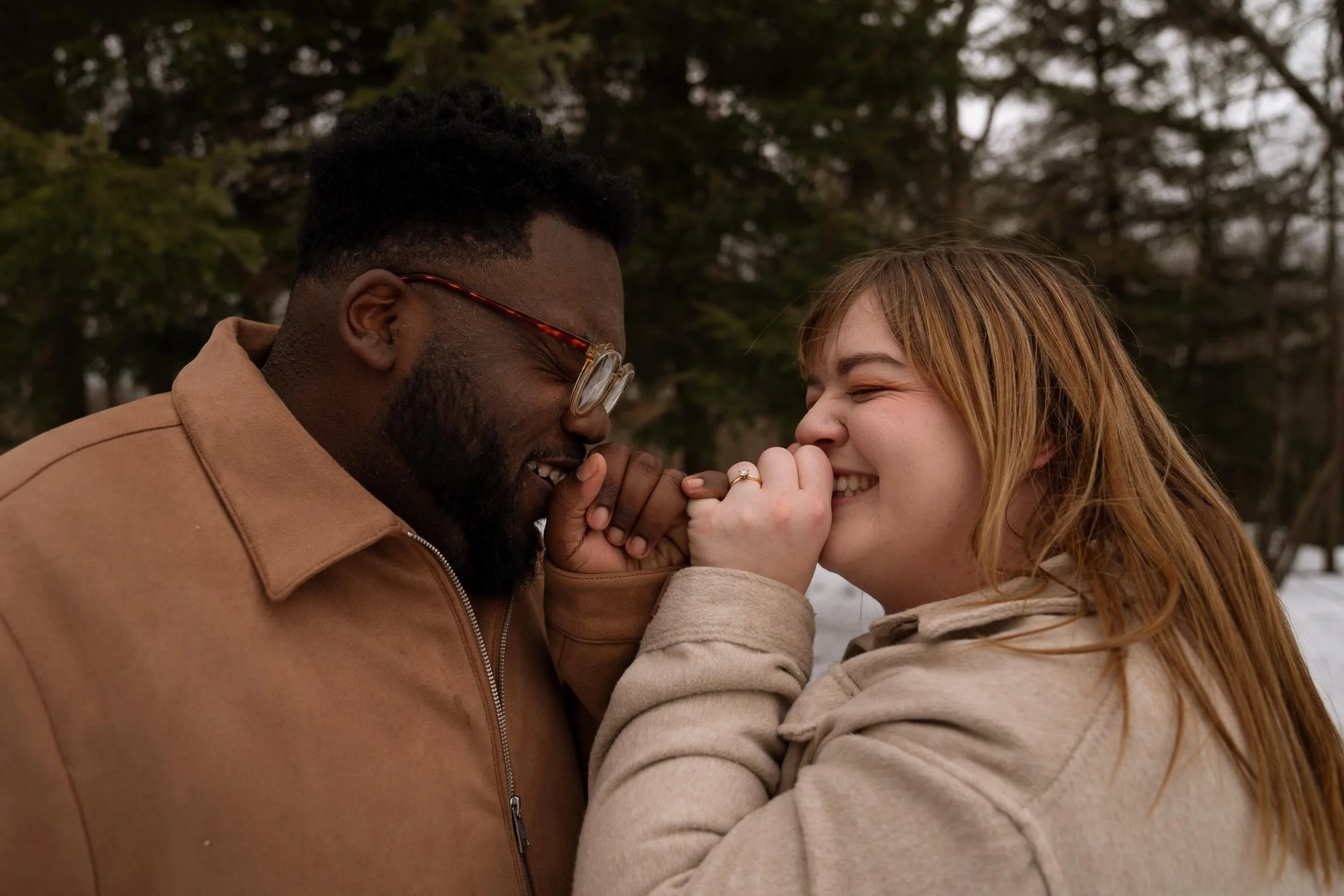 A man and a woman are smiling and holding hands closely, with the woman’s hand showing an engagement ring. They are outdoors, surrounded by trees.