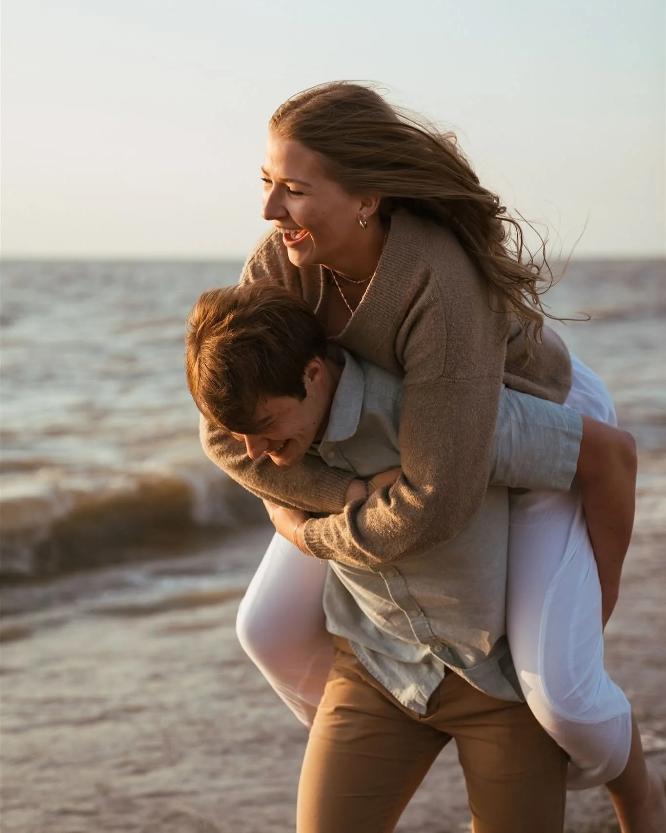 A woman with long, wavy blonde hair smiling while giving a piggyback ride to a man at the beach, with the ocean and sky in the background.