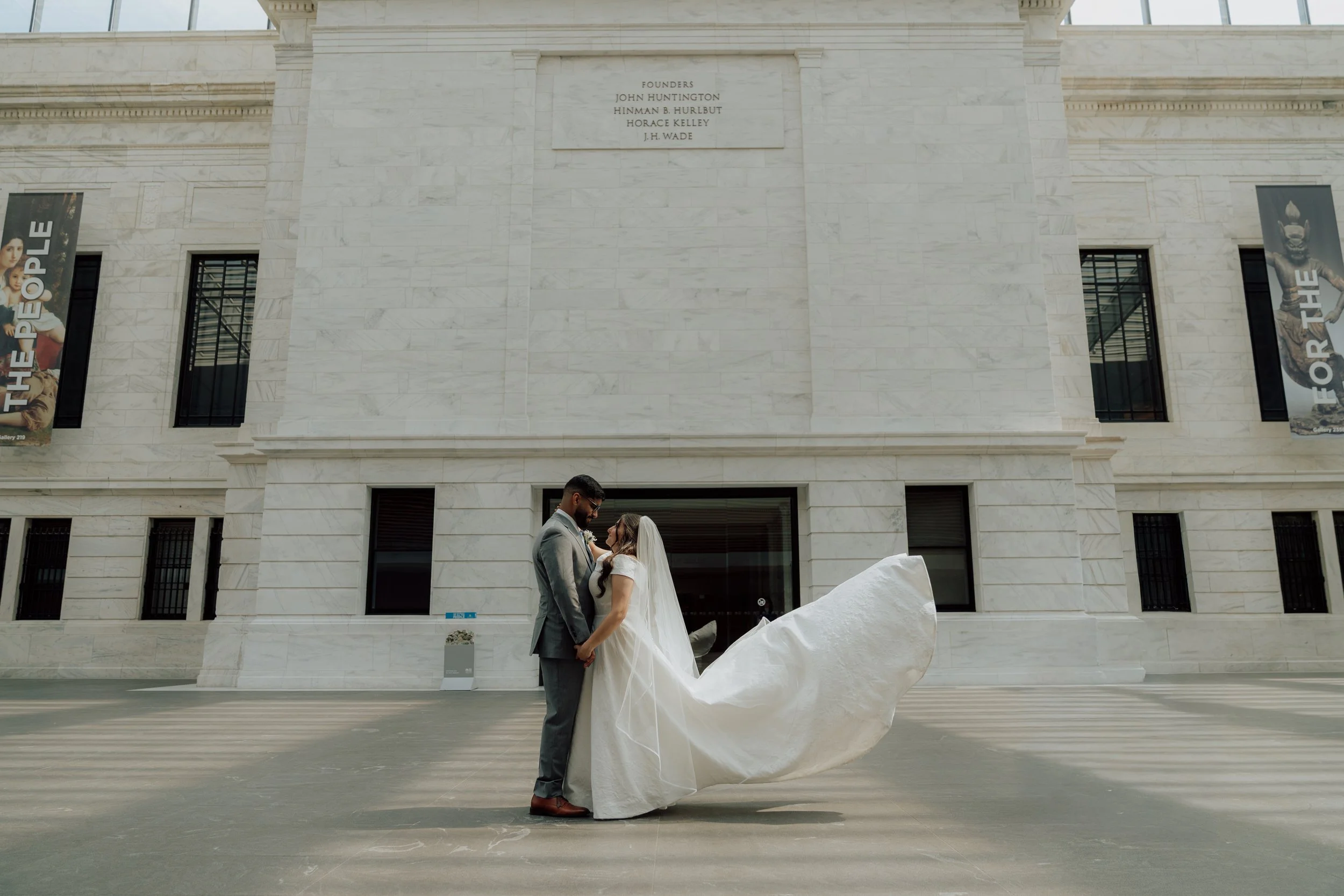 A bride and groom holding hands in front of a large white building with marble walls, with banners on either side and a plaque above, during a wedding photoshoot.