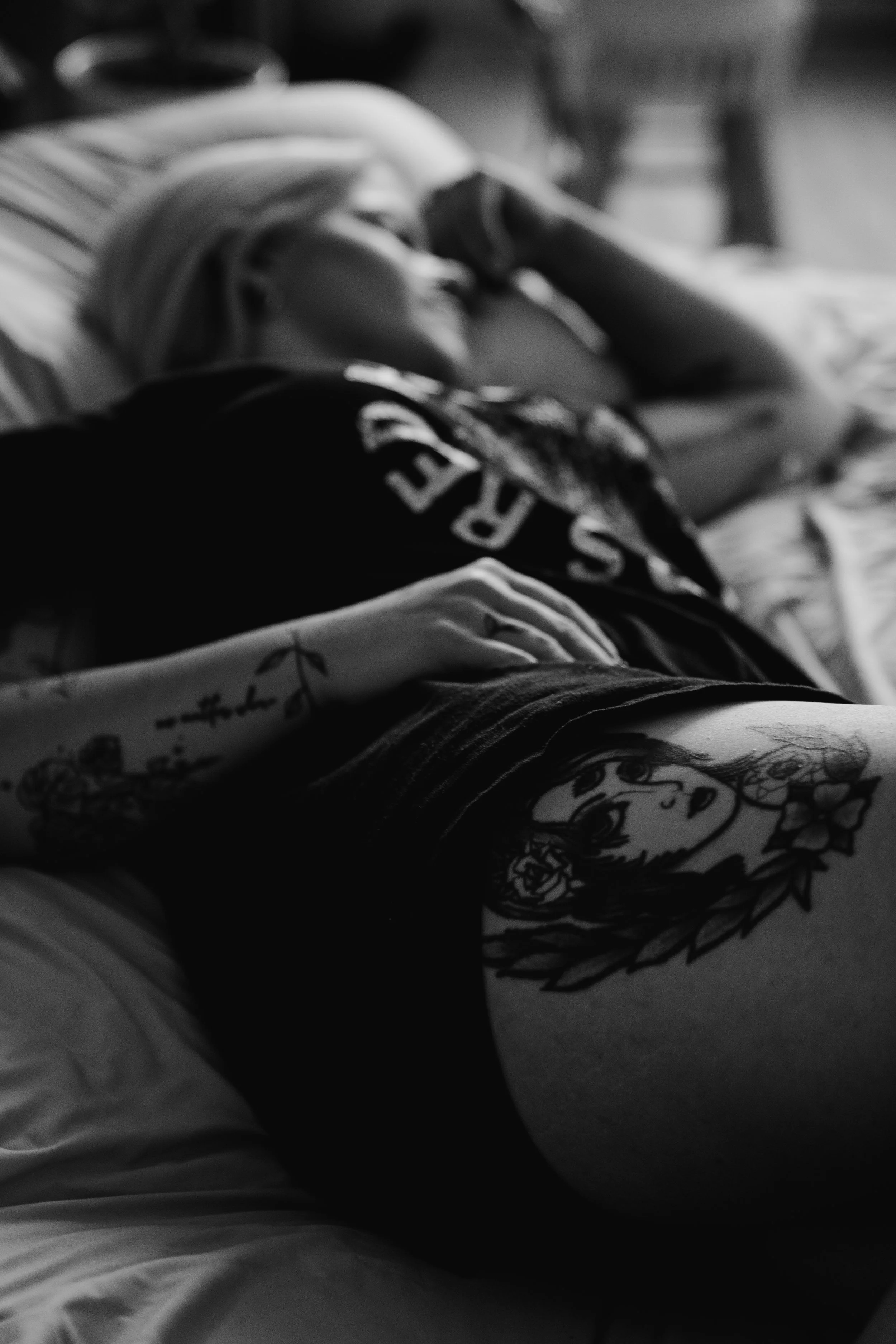 Black and white photo of a woman with tattoos, lying down with her head resting on her arm and smiling, surrounded by patterned fabrics.