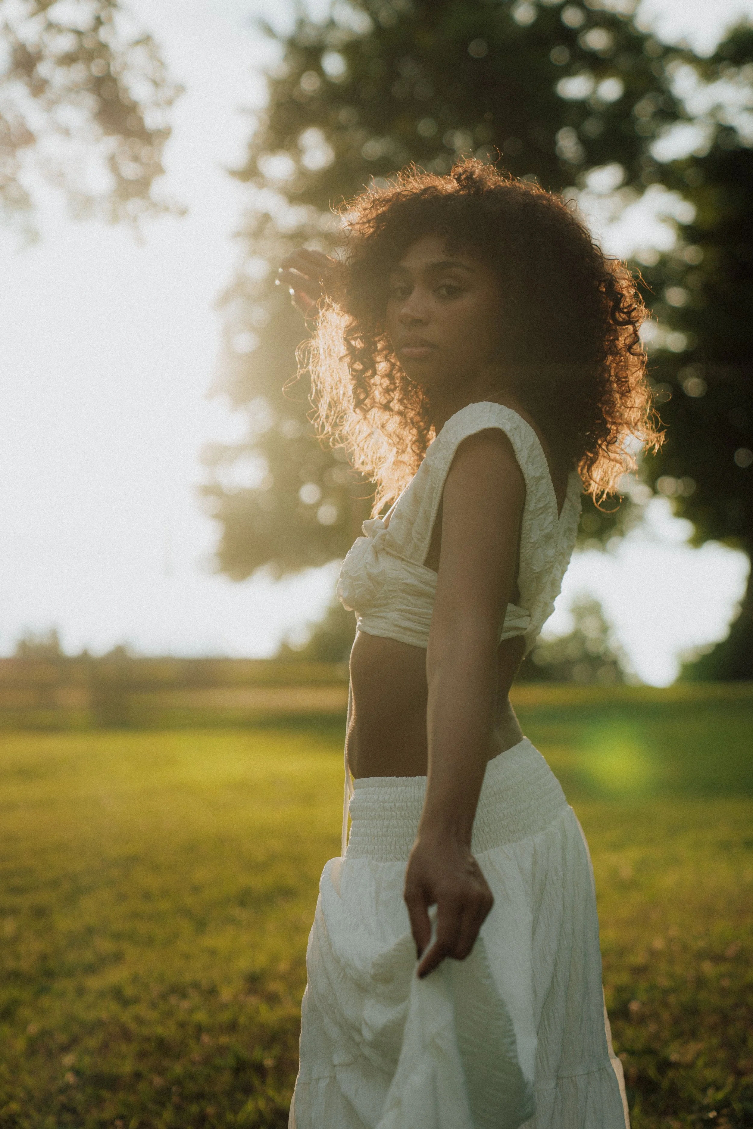 A woman with curly hair standing outdoors in a grassy area during sunlight, wearing a white sleeveless top and white pants, looking at the camera.