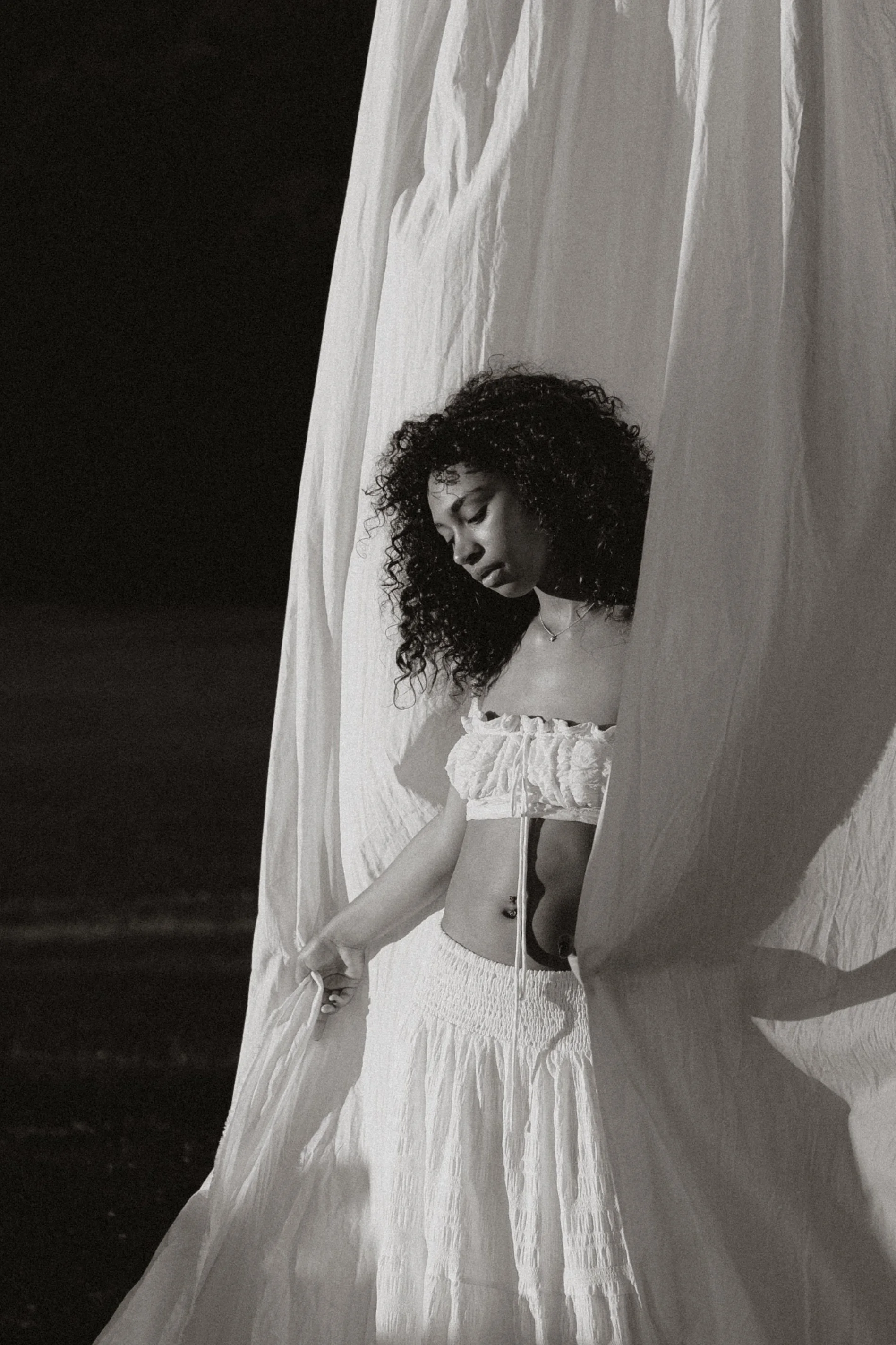 A young woman with curly hair sitting on a white bed, wearing a white crop top and skirt, looking down with closed eyes, surrounded by white curtains.