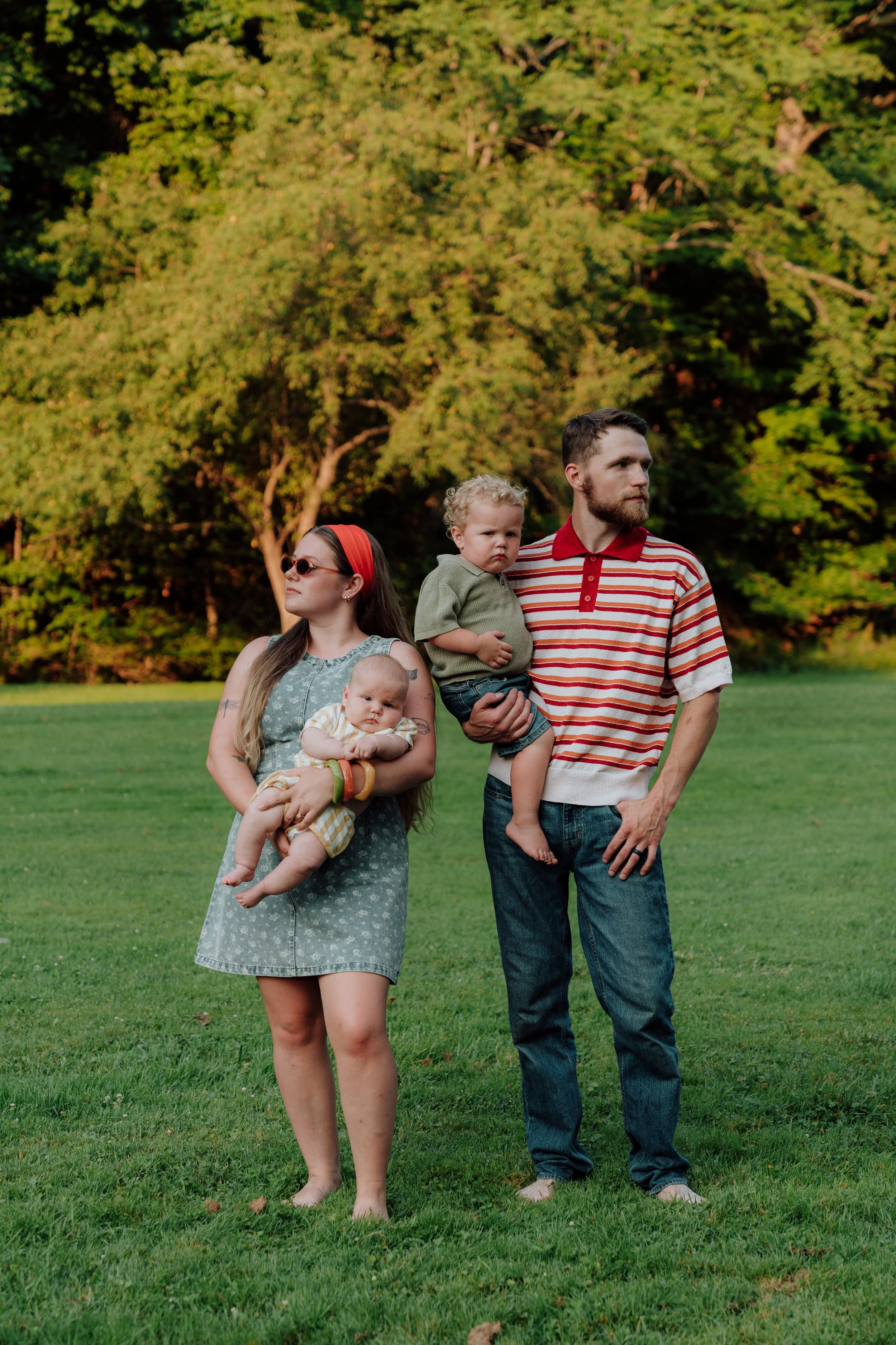 A family of four standing outdoors on a grassy field with trees in the background. The woman has long hair, sunglasses, and a sleeveless dress, holding a baby. The man has a beard, wearing a striped shirt, and is holding a young boy. The woman is loo