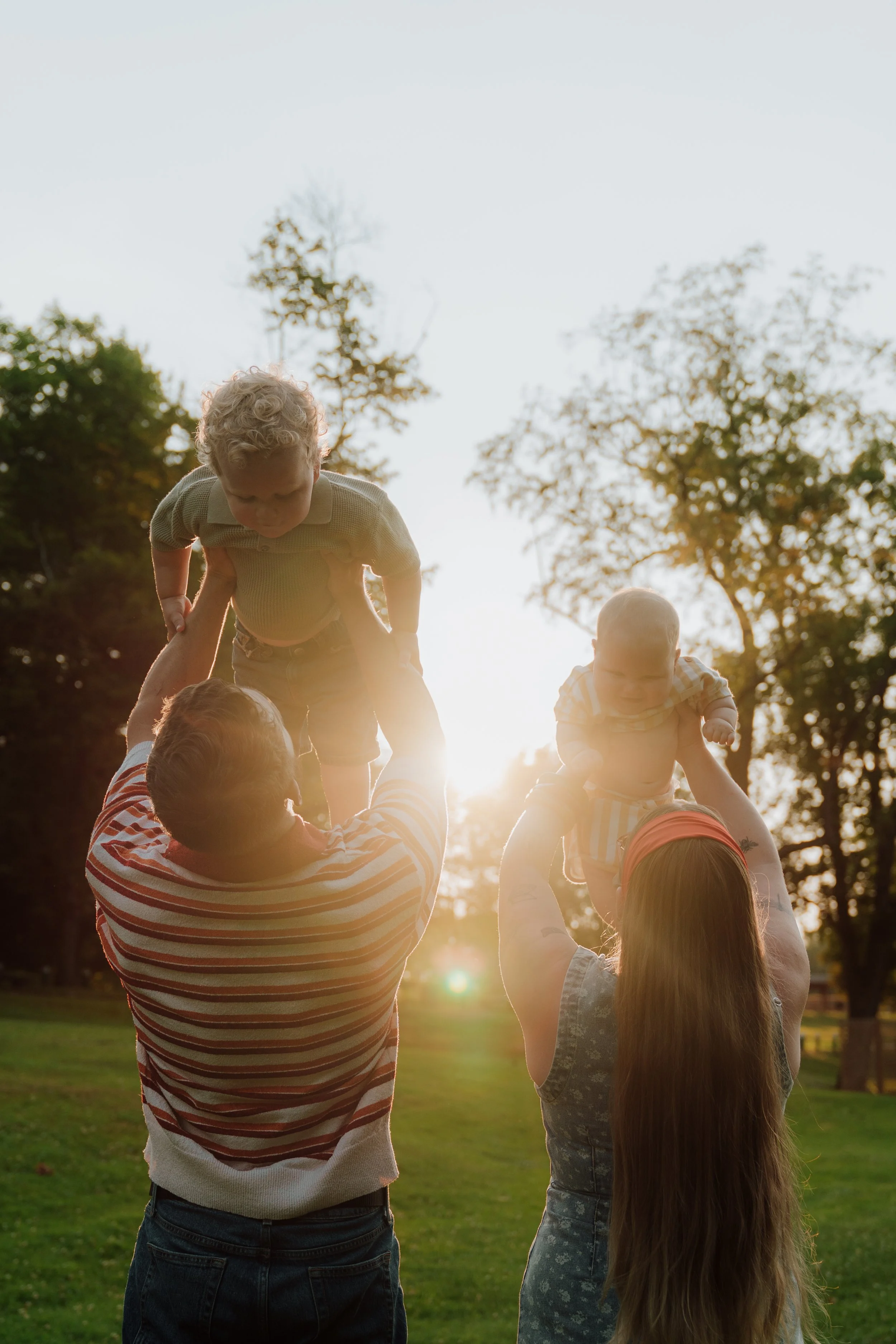 People forming a human pyramid outdoors during sunset, with children at the top and an adult at the bottom, surrounded by trees.