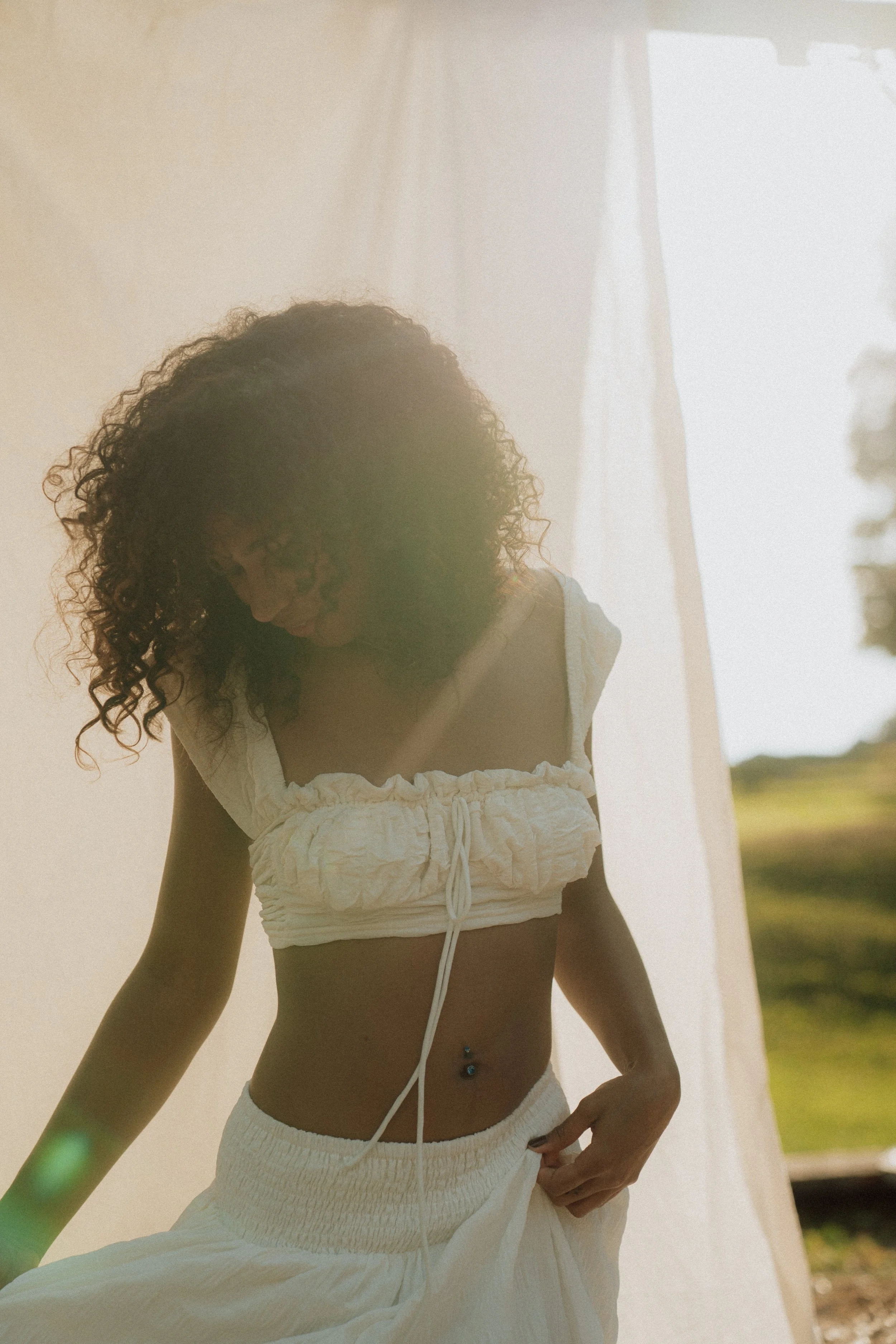 A woman with curly hair wearing a white crop top and white pants, standing outdoors near a light-colored wall, with a field and trees in the background.