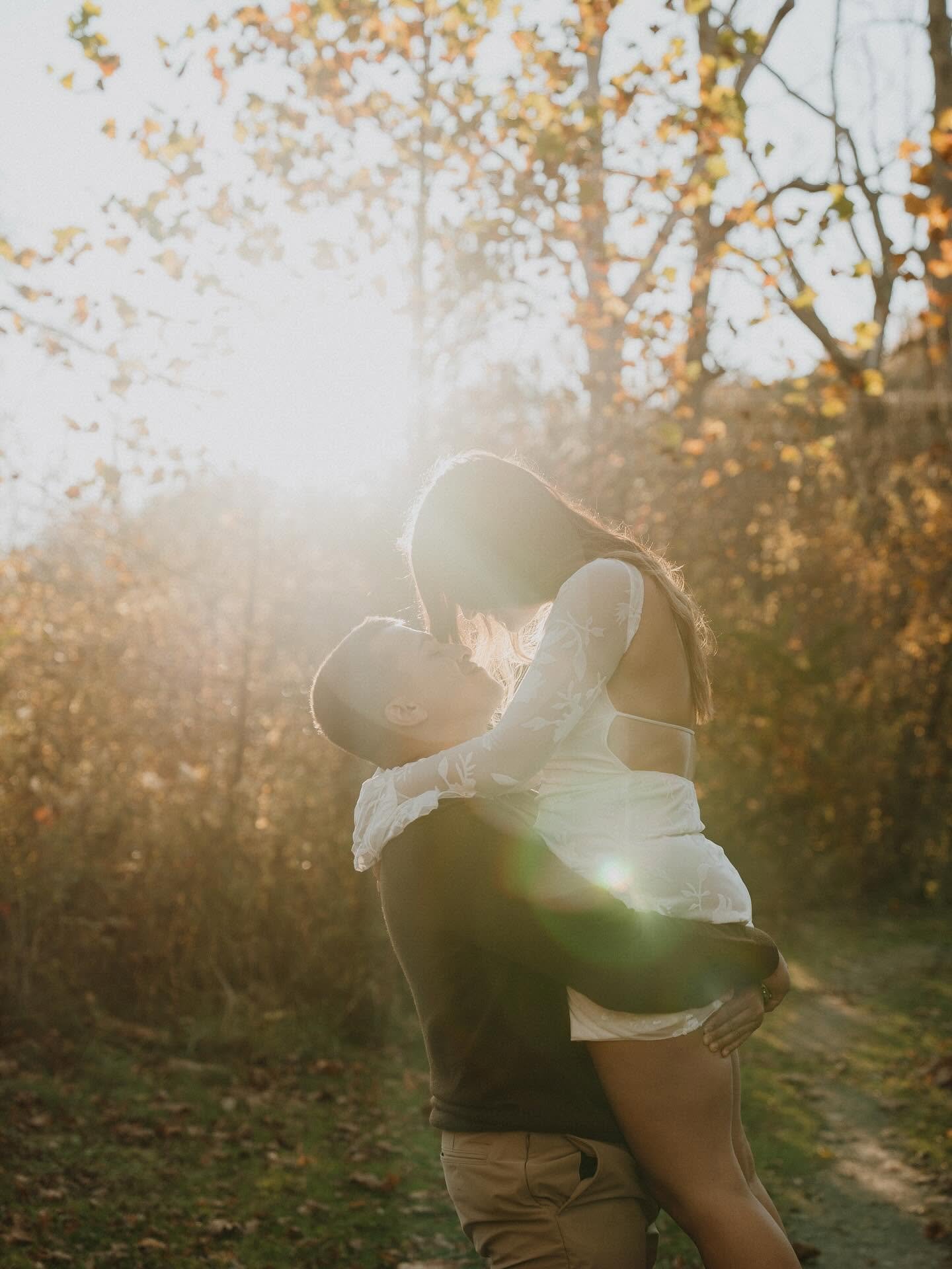 A couple sharing a kiss outdoors in a park with fall foliage in the background, backlit by sunlight.