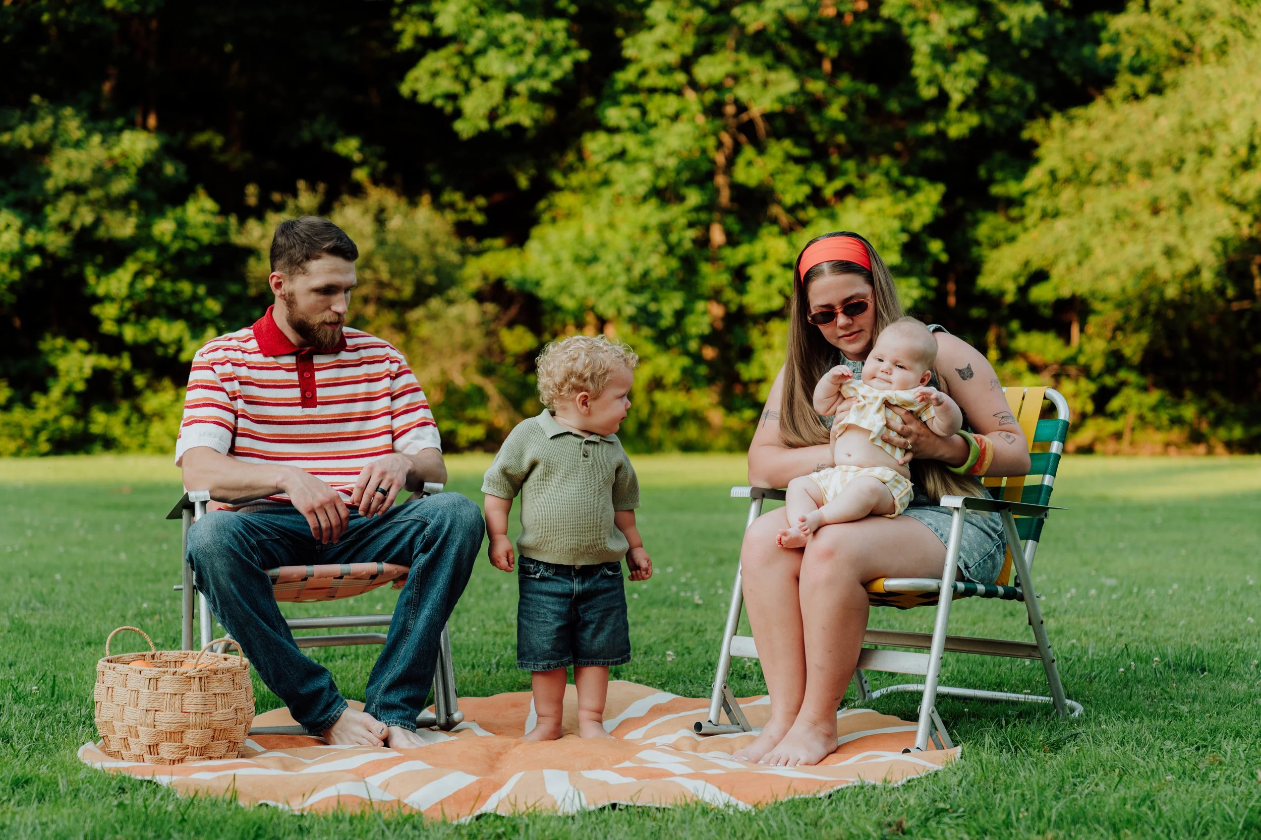 Family enjoying a picnic outdoors on a sunny day, with two adults and two children sitting on a blanket and chairs on a grassy field surrounded by trees