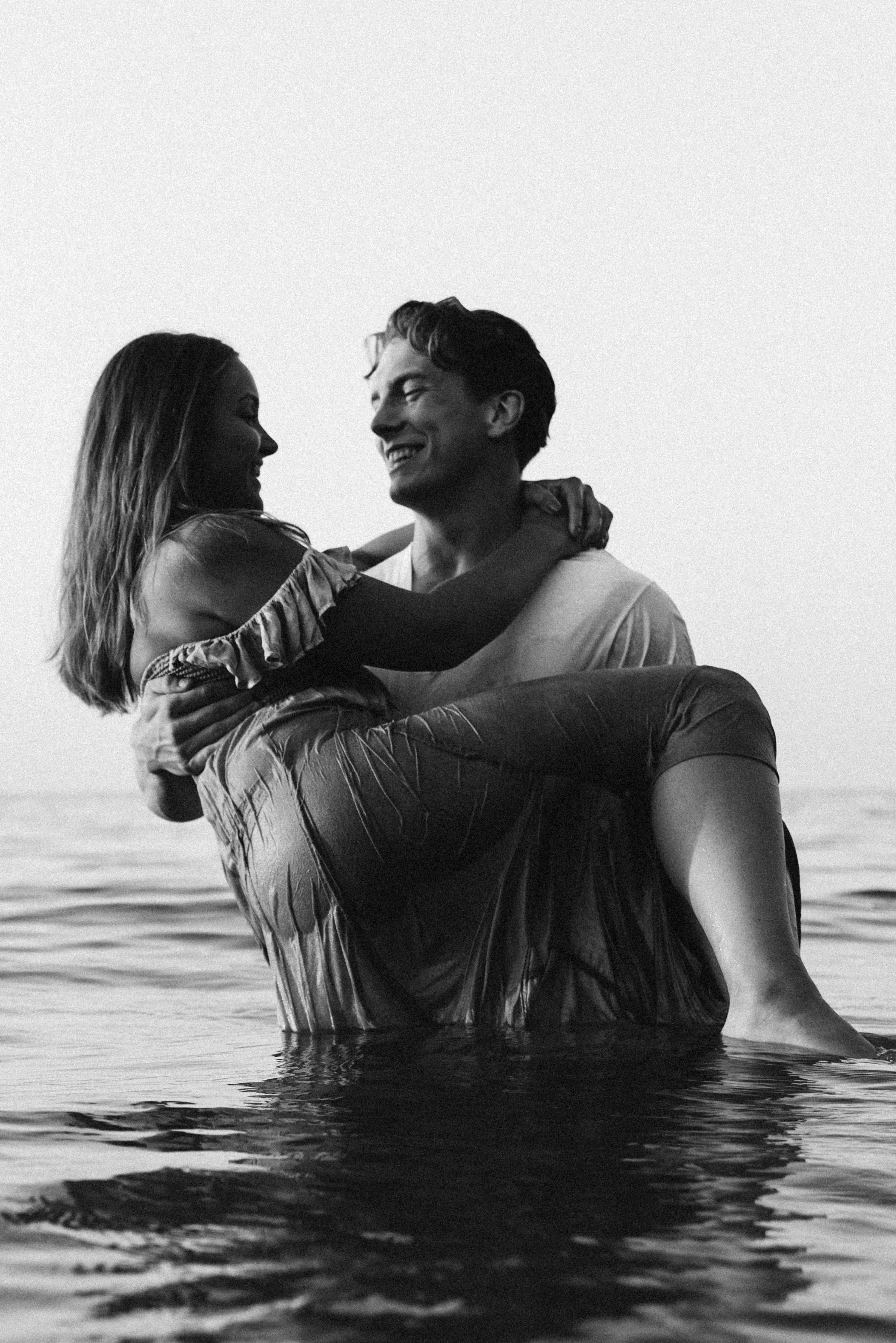 A couple in the water, with the woman being carried by the man, both smiling and appearing happy, at the beach during sunset or sunrise.