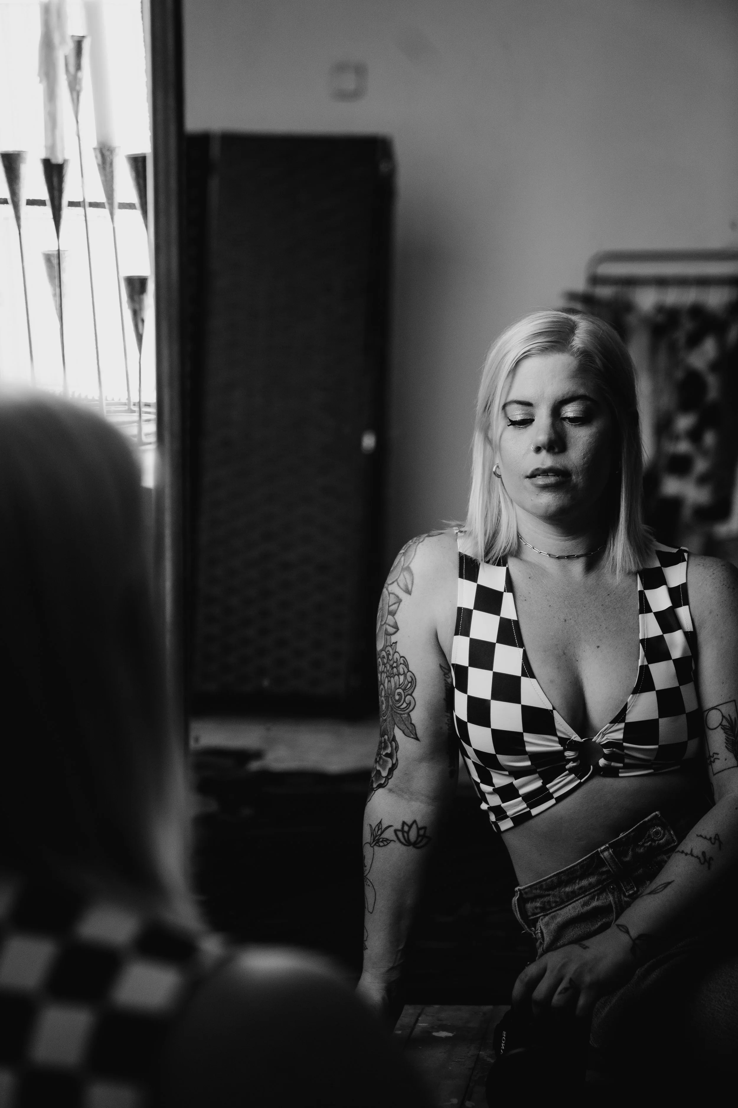 Black and white photo of a woman with tattoos on her arm, wearing a checkered top, sitting on the floor and looking down, with a mirror reflecting her face. The background includes a window with blinds and a wall with a small square object.