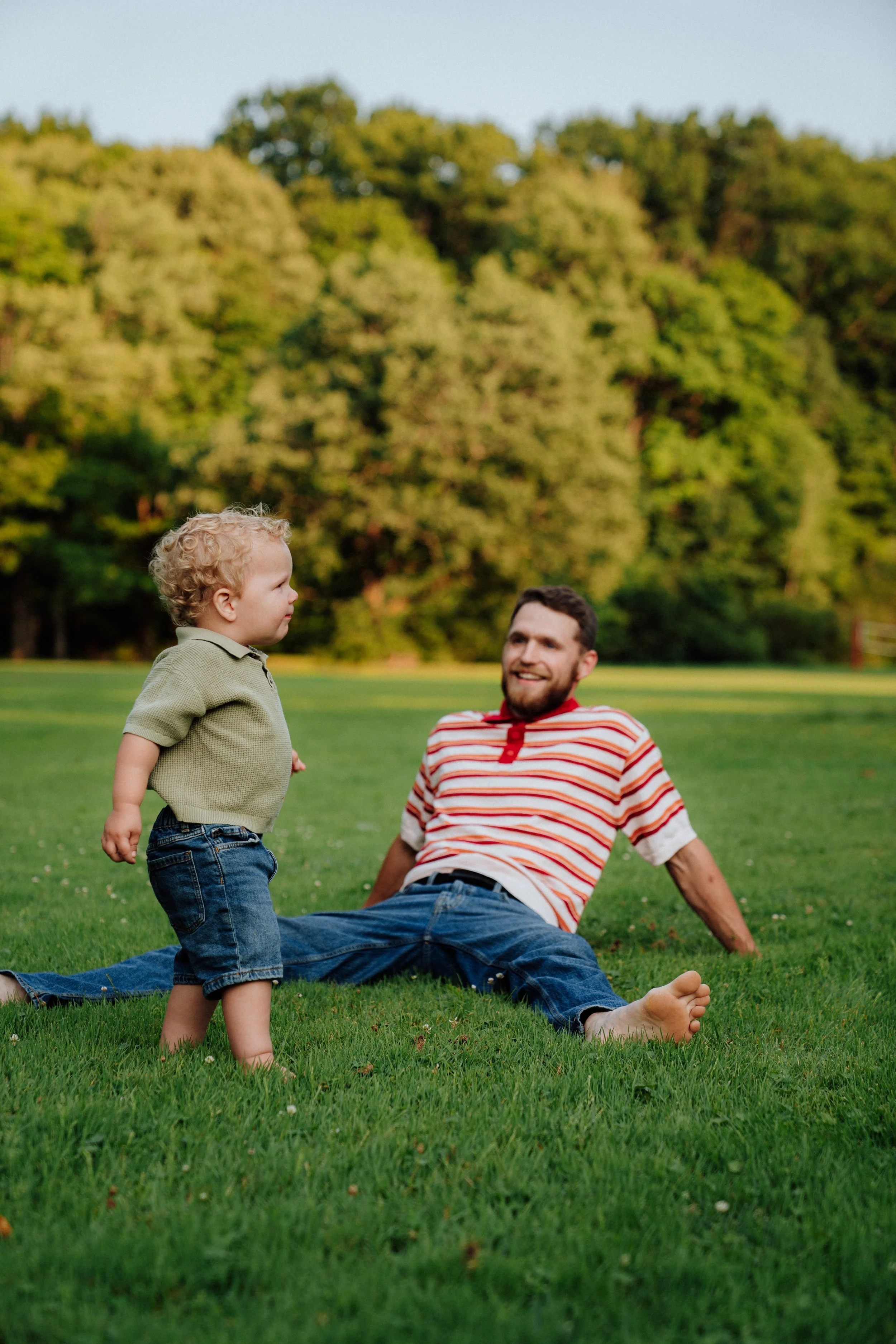 A father and his young son playing on a grassy field in the park, with trees in the background; the father is sitting on the ground smiling at the son who is standing and facing him.