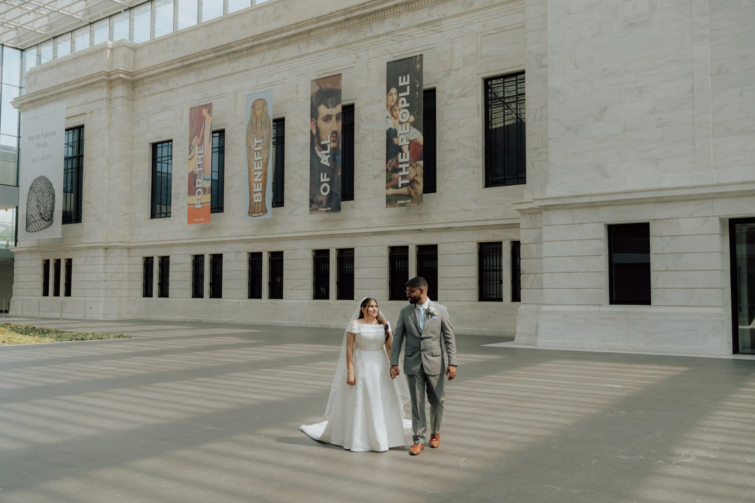 A bride and groom walking hand in hand outside a museum with banners displaying artwork and the text 'To the People of the All of the People.'