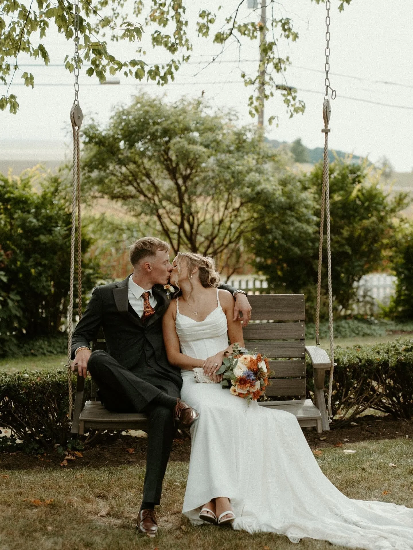 A newlywed couple sharing a kiss on a wooden swing in a garden setting during daytime.