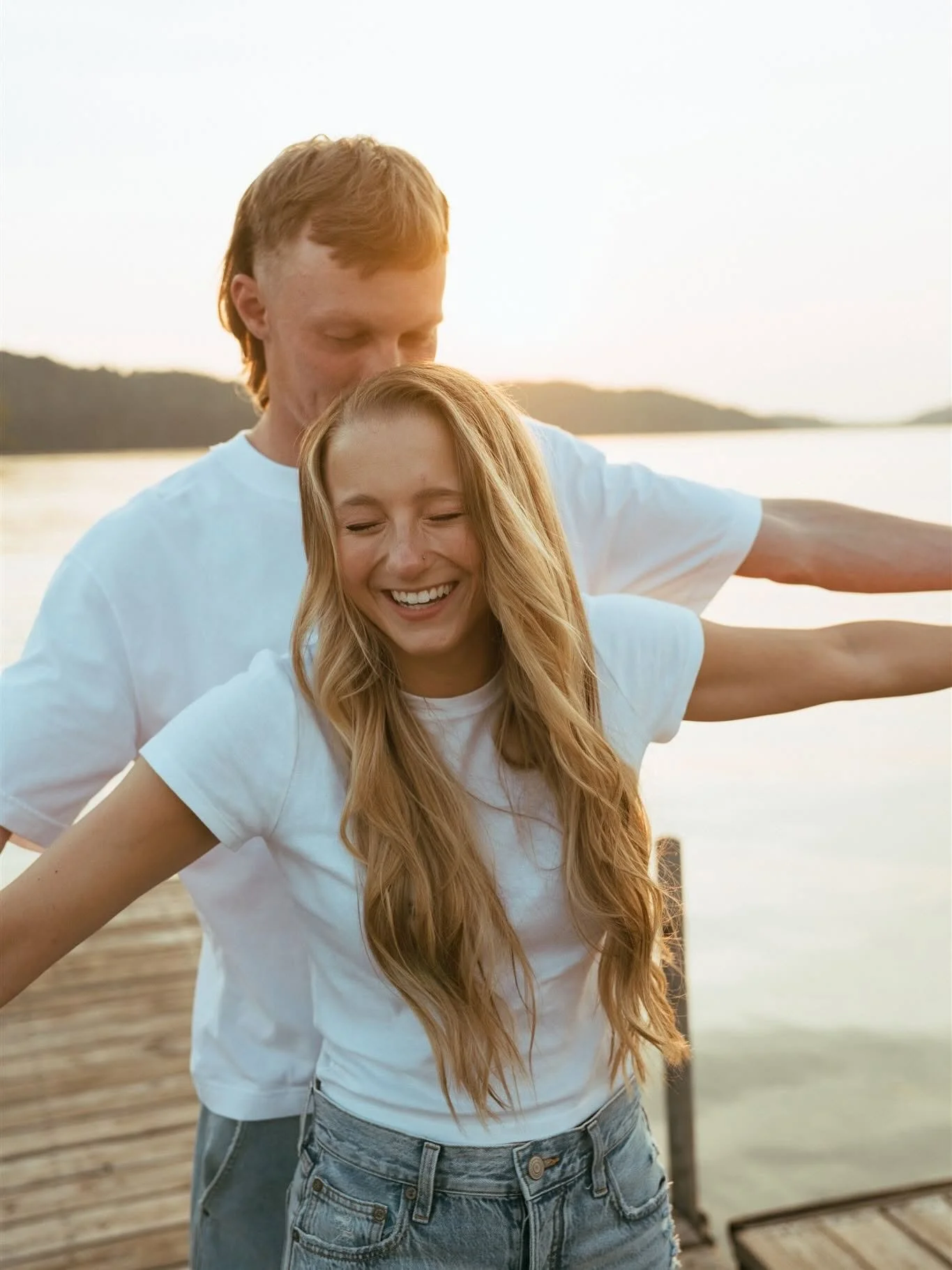 A young woman with long blonde hair and a young man with red hair are outdoors near a body of water during sunset. The woman is smiling with her eyes closed, and the man is standing behind her with his arms outstretched, also smiling. They are both w