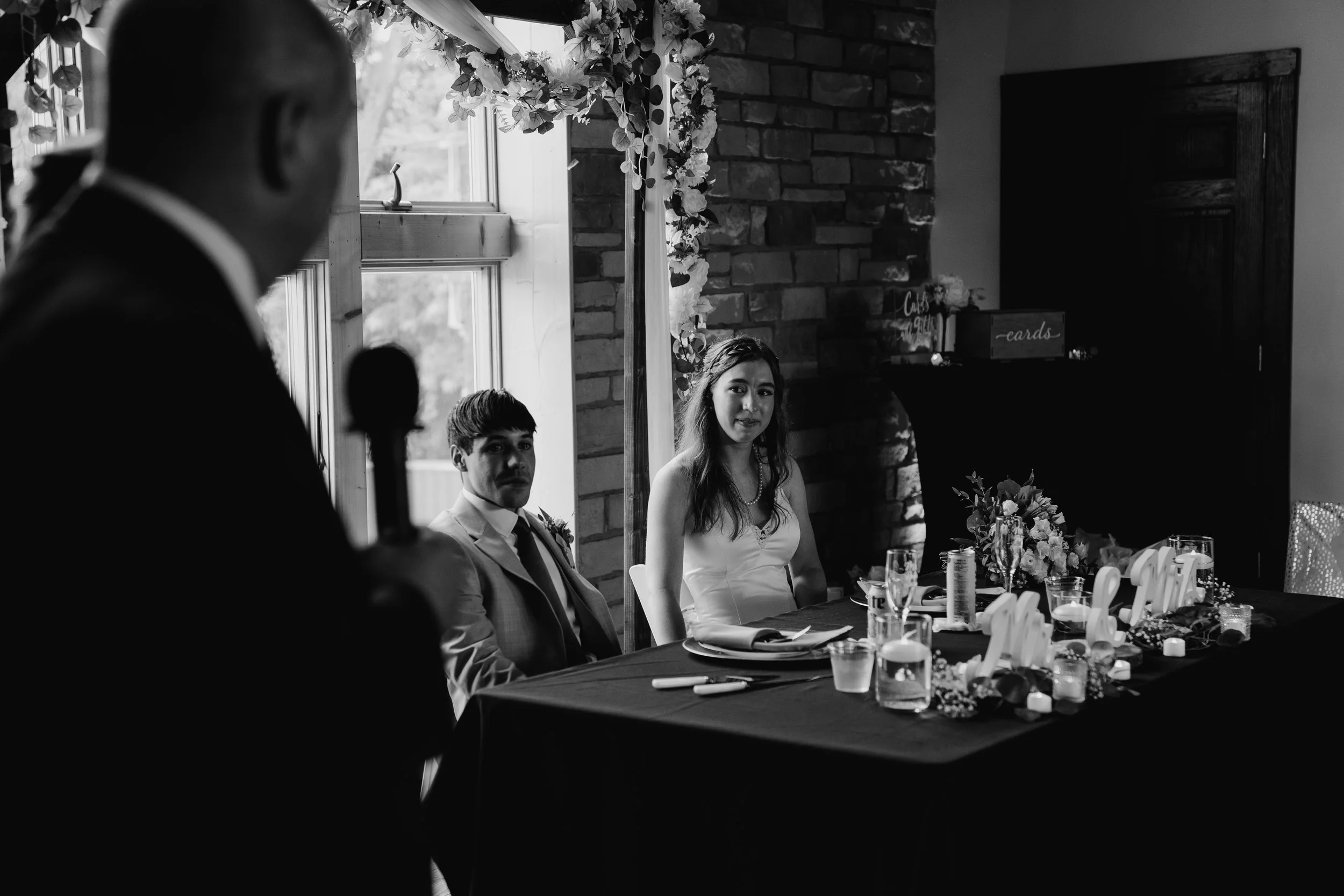 Black and white photo of a wedding reception with a bride and groom seated at a decorated table, while a man is speaking at a microphone in the foreground, and floral decorations are visible near the window.
