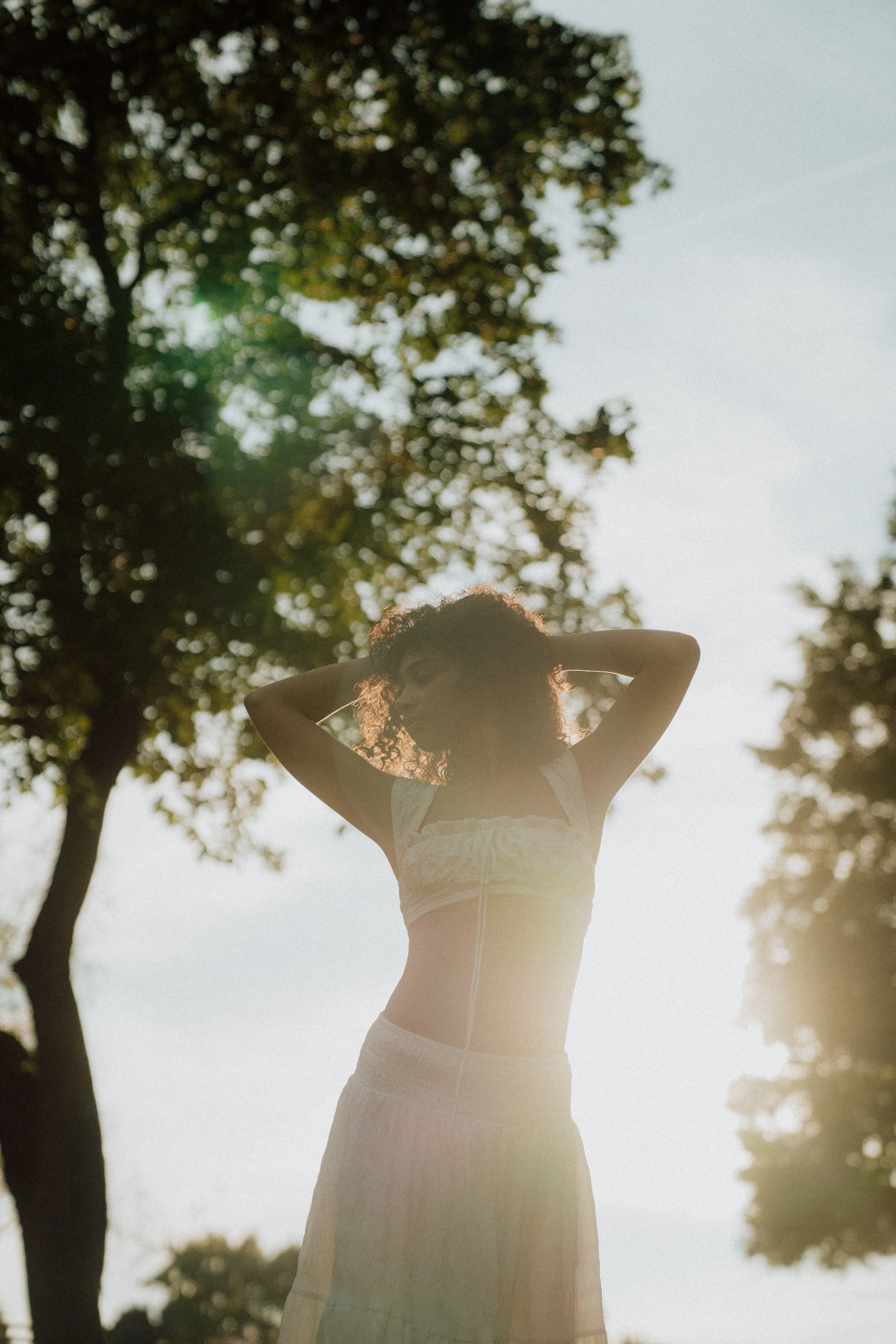 A woman with curly hair standing outdoors with sunlight shining through the trees, wearing a light-colored crop top and a long skirt, with her arms raised and hands behind her head.