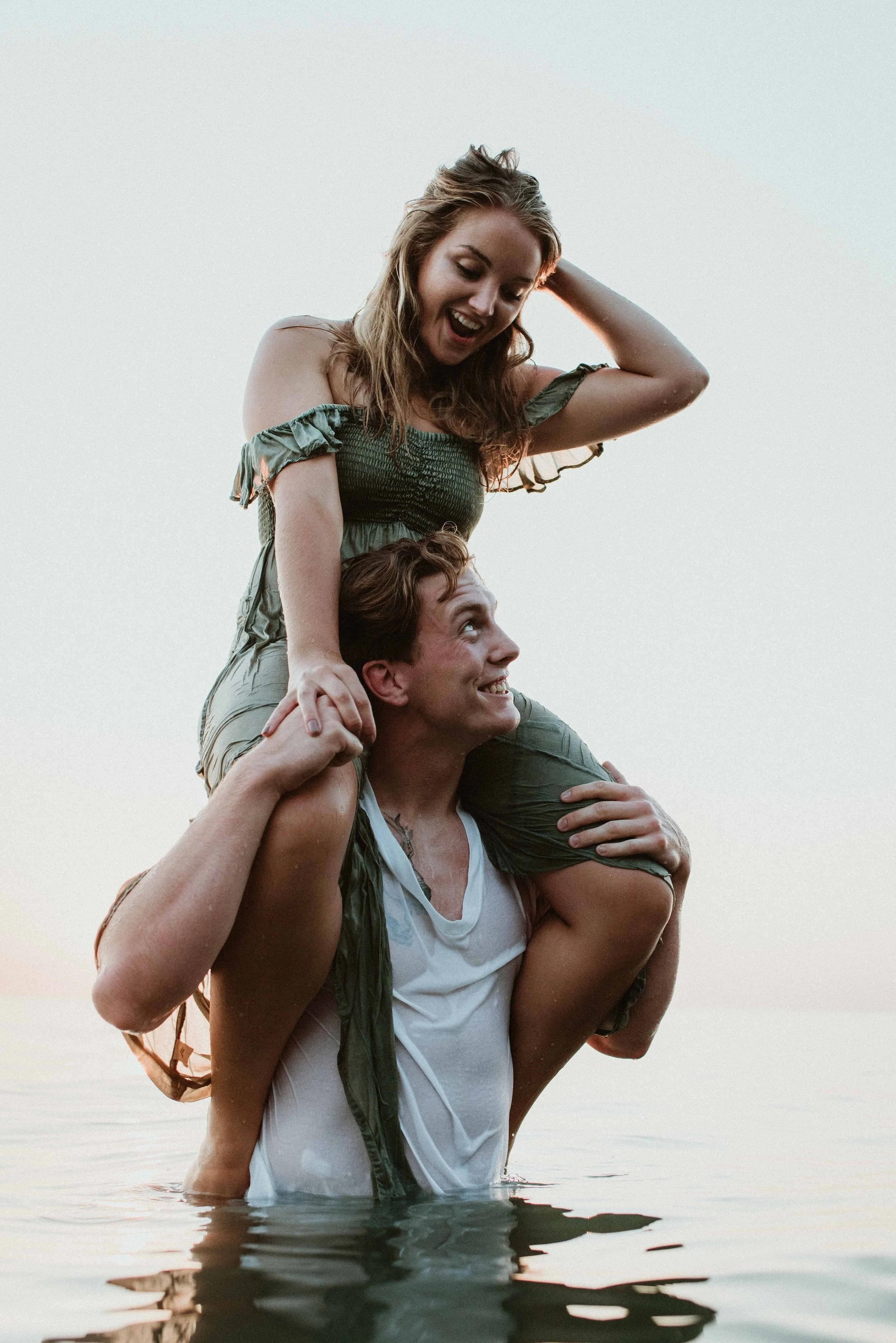 A man and woman are playing on the water, with the woman sitting on the man's shoulders. They are smiling and appear happy, enjoying a moment of fun together during sunset or sunrise.