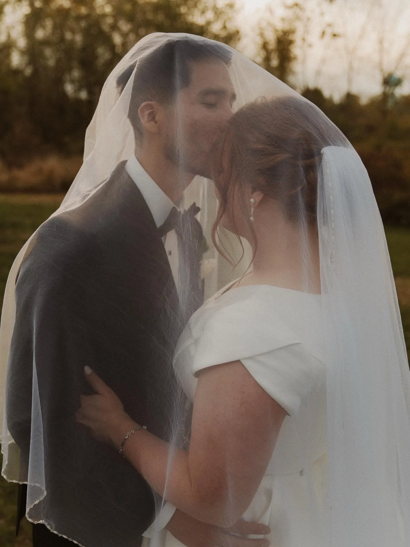 A bride and groom sharing a kiss, with the groom wearing a tuxedo and the bride in a wedding dress, under a bridal veil in an outdoor setting at sunset.