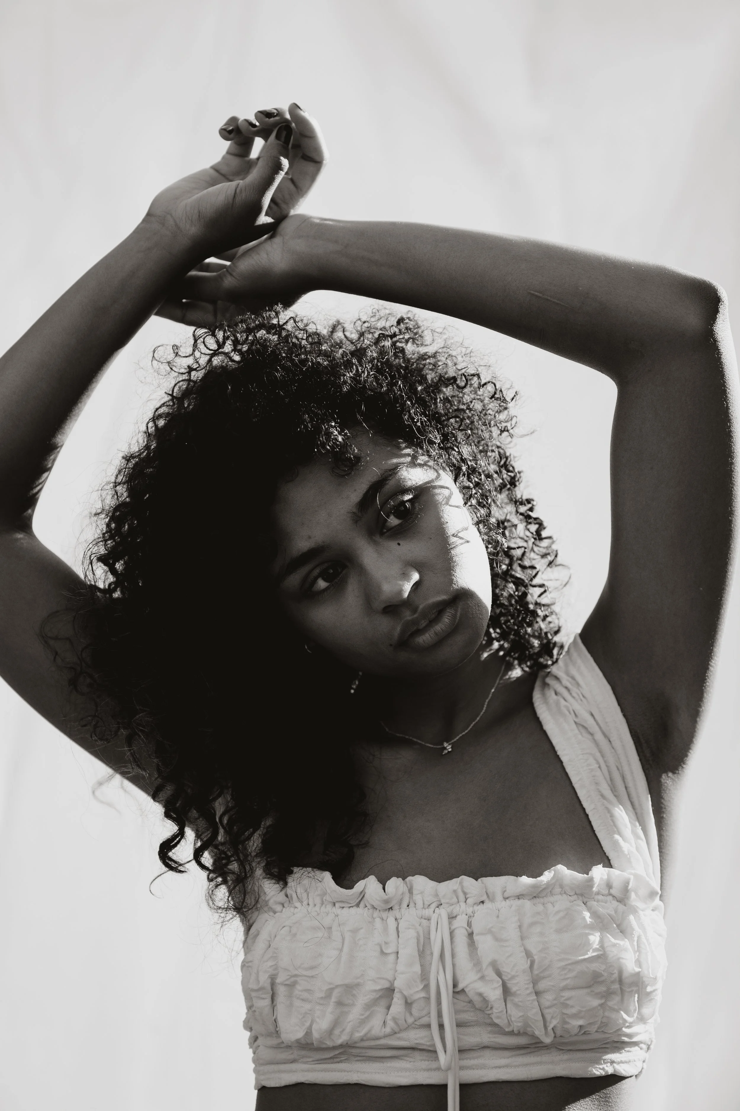 A black-and-white photo of a woman with curly hair, wearing a sleeveless top, posing with arms raised above her head and hands intertwined, looking directly at the camera.