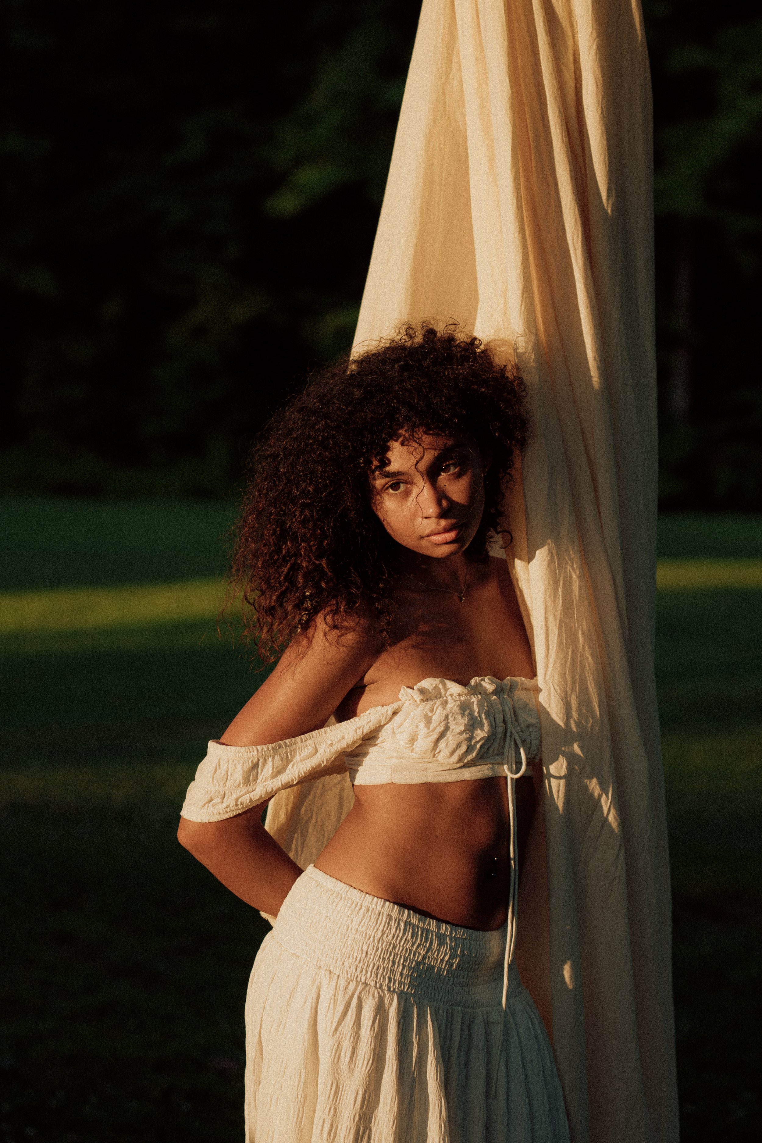 A woman with curly hair standing outdoors near a large beige fabric, wearing a cream-colored off-the-shoulder top and matching skirt, with the sun casting a warm glow.