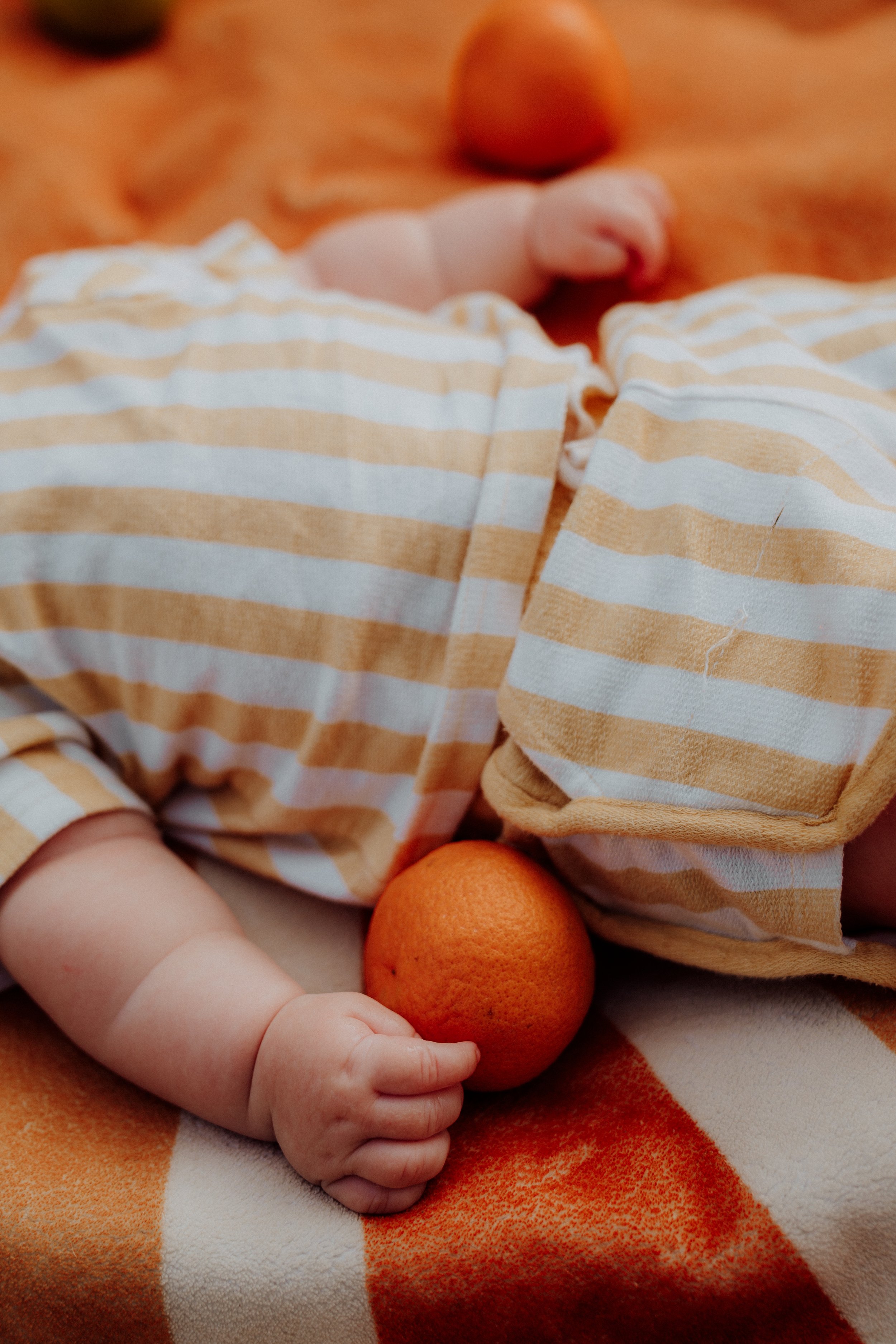 Close-up of a baby dressed in a yellow and white striped shirt, holding an orange. The baby has a chubby arm and a hand gripping the orange, with other oranges visible in the background.
