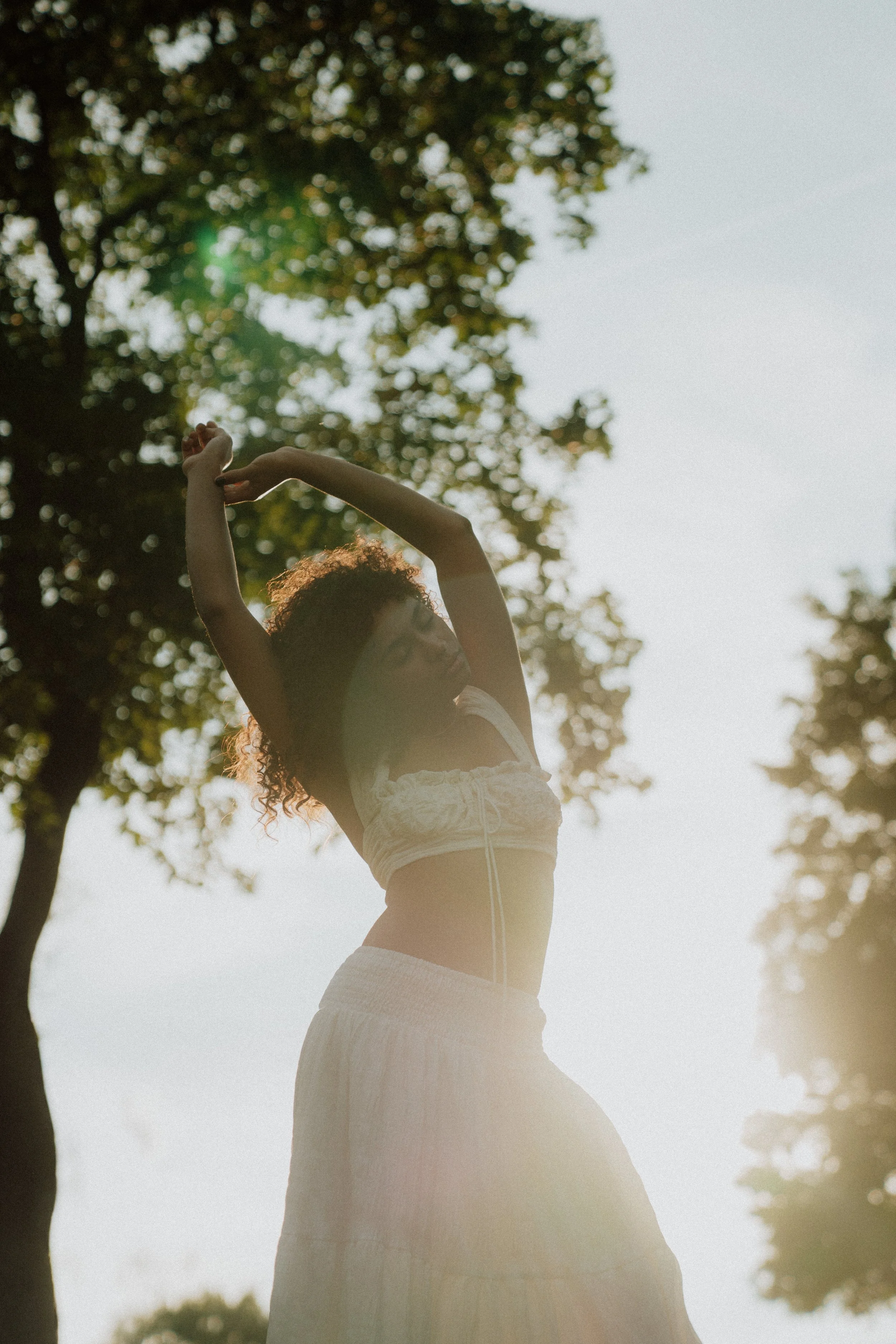 A woman with curly hair wearing glasses, a white crop top, and a long skirt, stretching outdoors with trees in the background and sunlight filtering through the leaves.