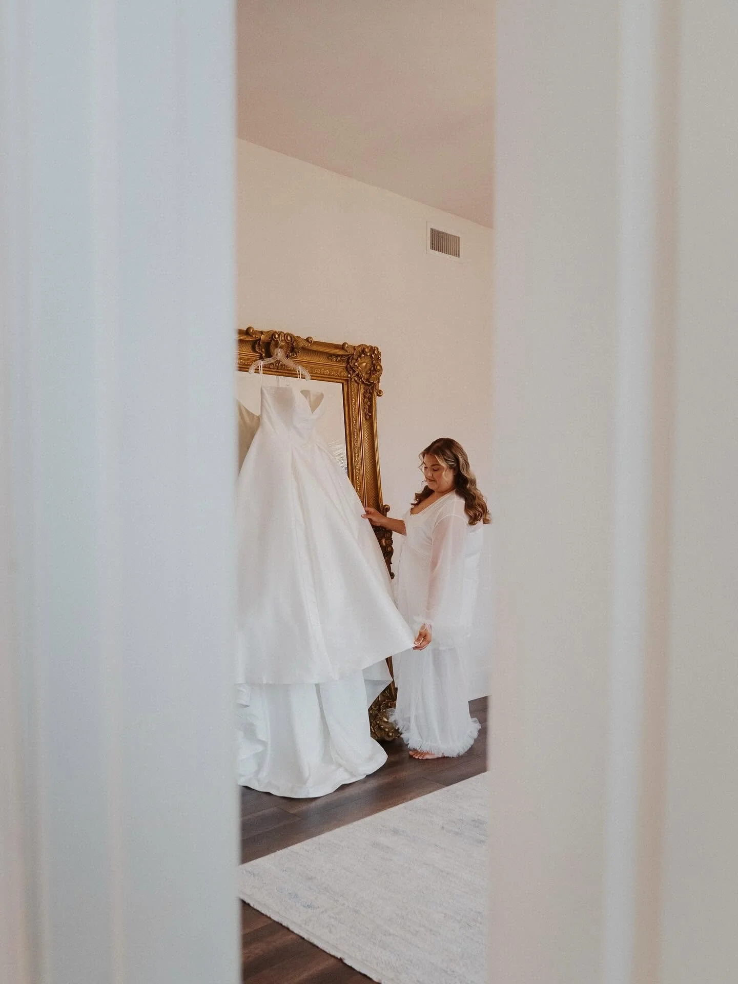 A woman in a white robe is looking at a white wedding dress hanging on a gold ornate mirror frame, seen through a slightly open door.