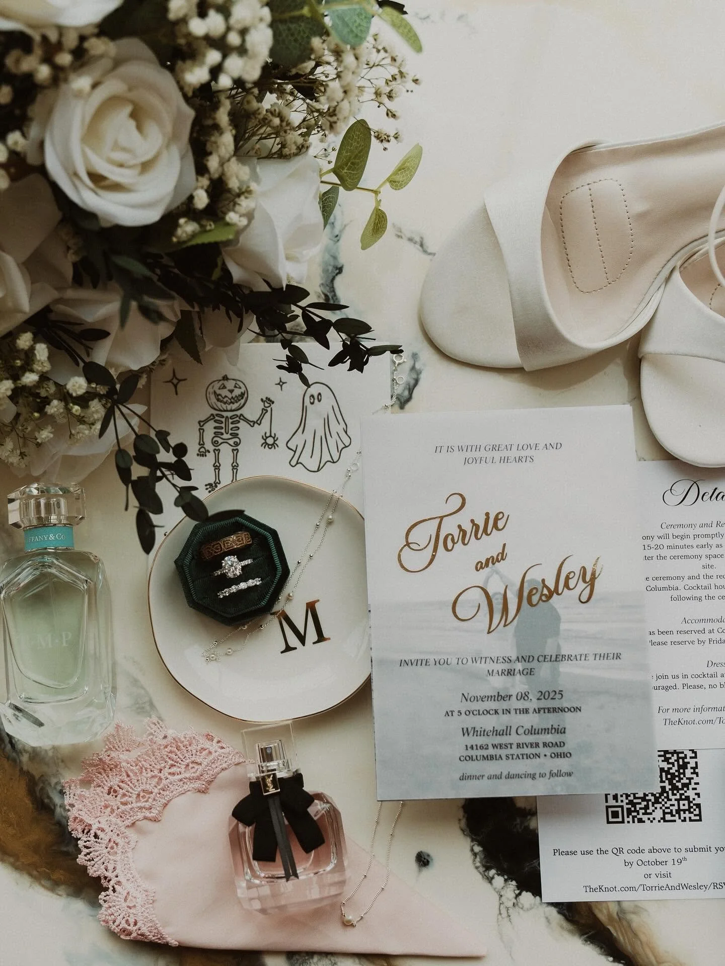 Wedding invitation for Torrie and Wesley with floral arrangement, jewelry, perfume, and wedding details scattered on a marble surface.