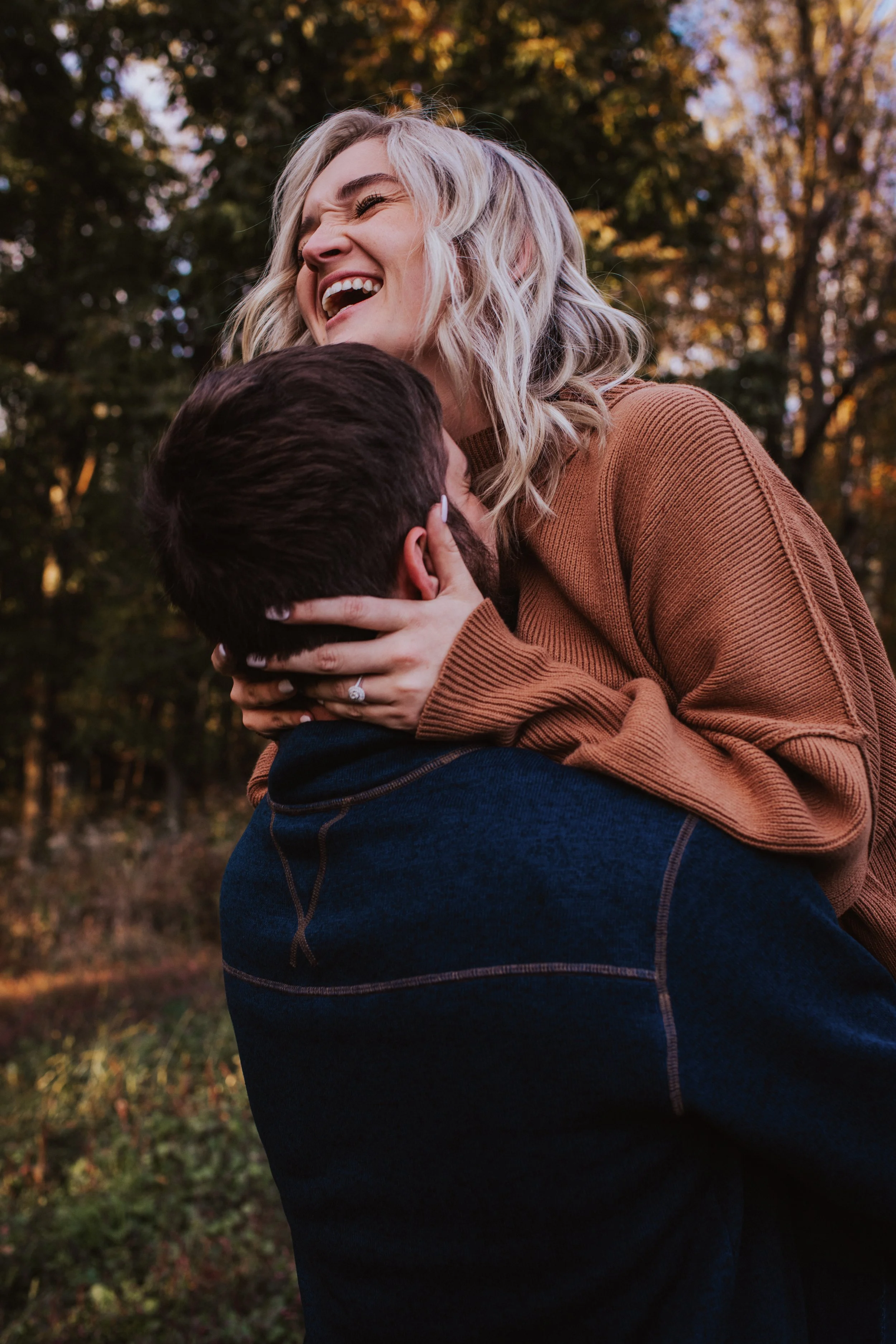 A woman with blonde wavy hair smiling and laughing while hugging a man in an outdoor setting with trees in the background during fall.