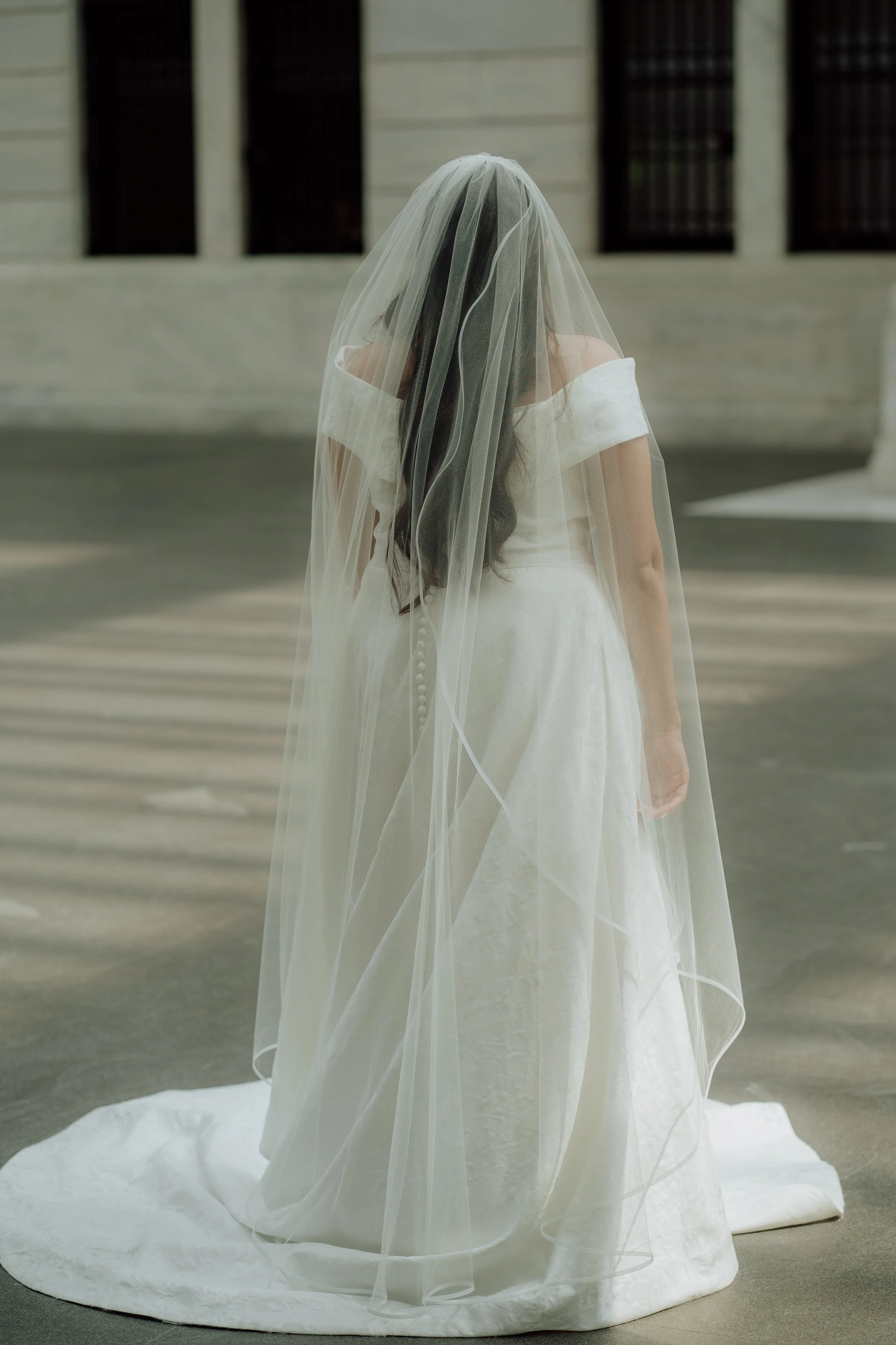 A woman in a wedding dress with a veil standing outdoors on a paved area in front of a building with barred windows.