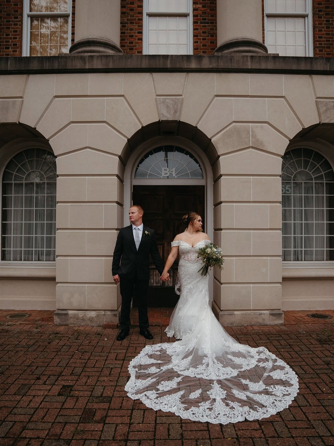 A bride and groom standing hand in hand outside a large building with brick walls and arched windows. The bride is wearing a white wedding gown with lace details and a long train, holding a bouquet, and the groom is in a black suit with a patterned t