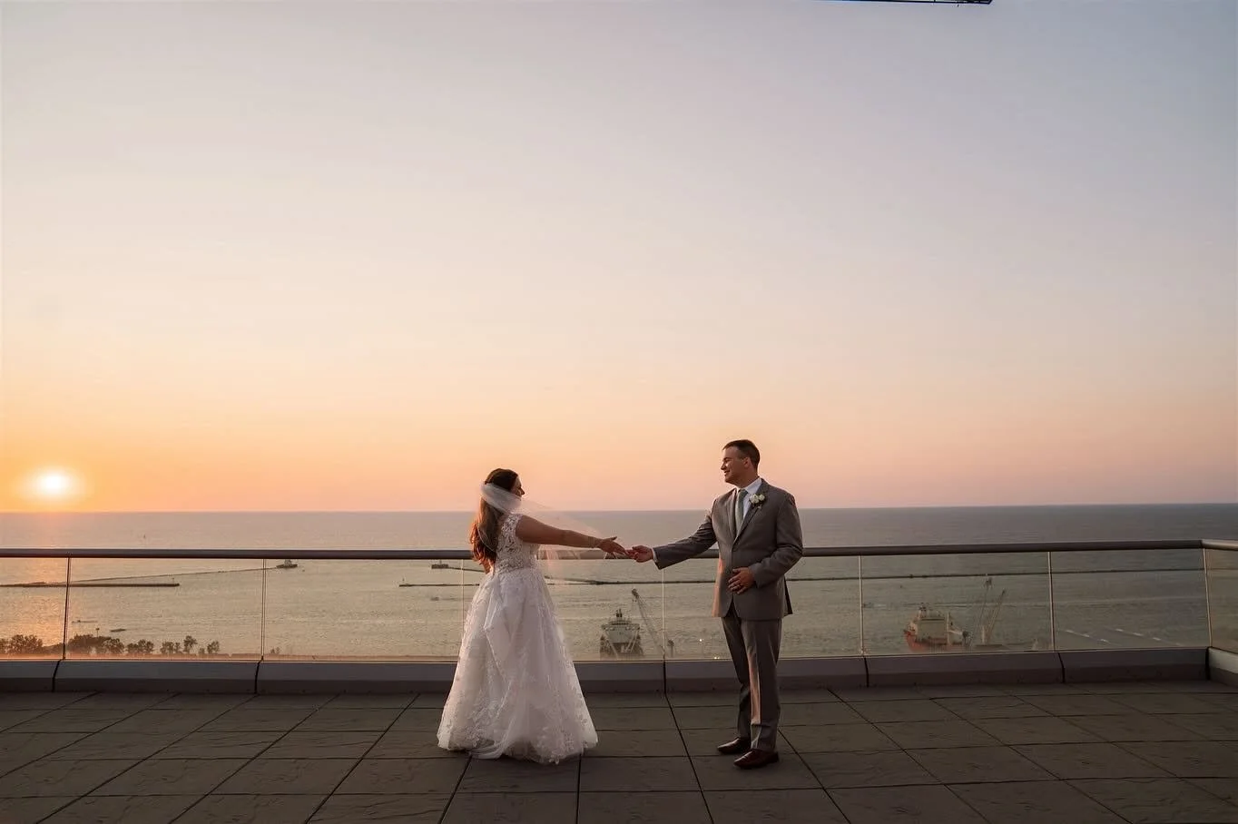 A bride and groom holding hands on a rooftop at sunset, with the ocean and ships in the background.