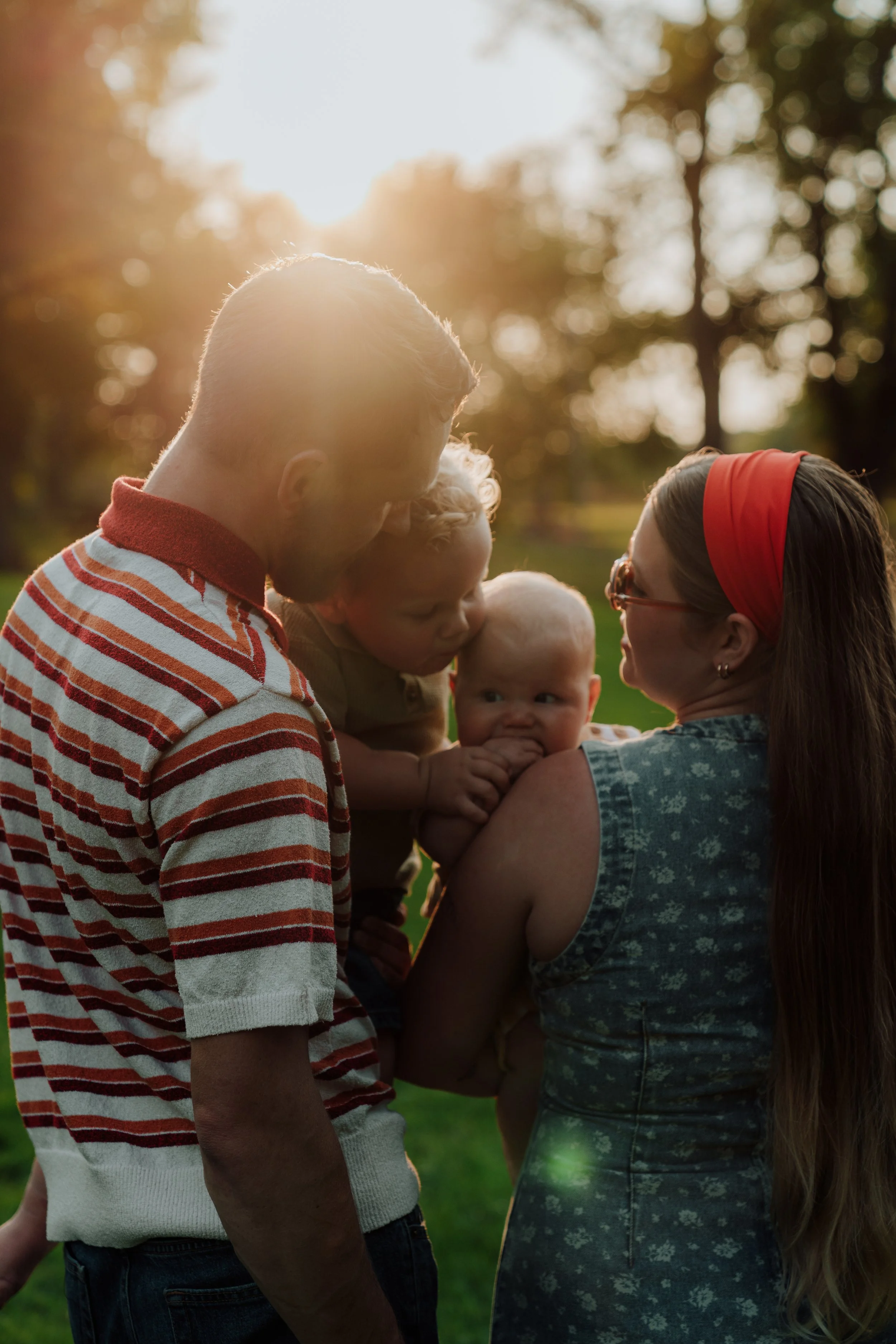 A family of four outdoors during golden hour, with warm sunlight illuminating their faces. The father, with short hair and a beard, wears a striped shirt. The mother wears sunglasses and a red headband. The baby, with a curious expression, is being h