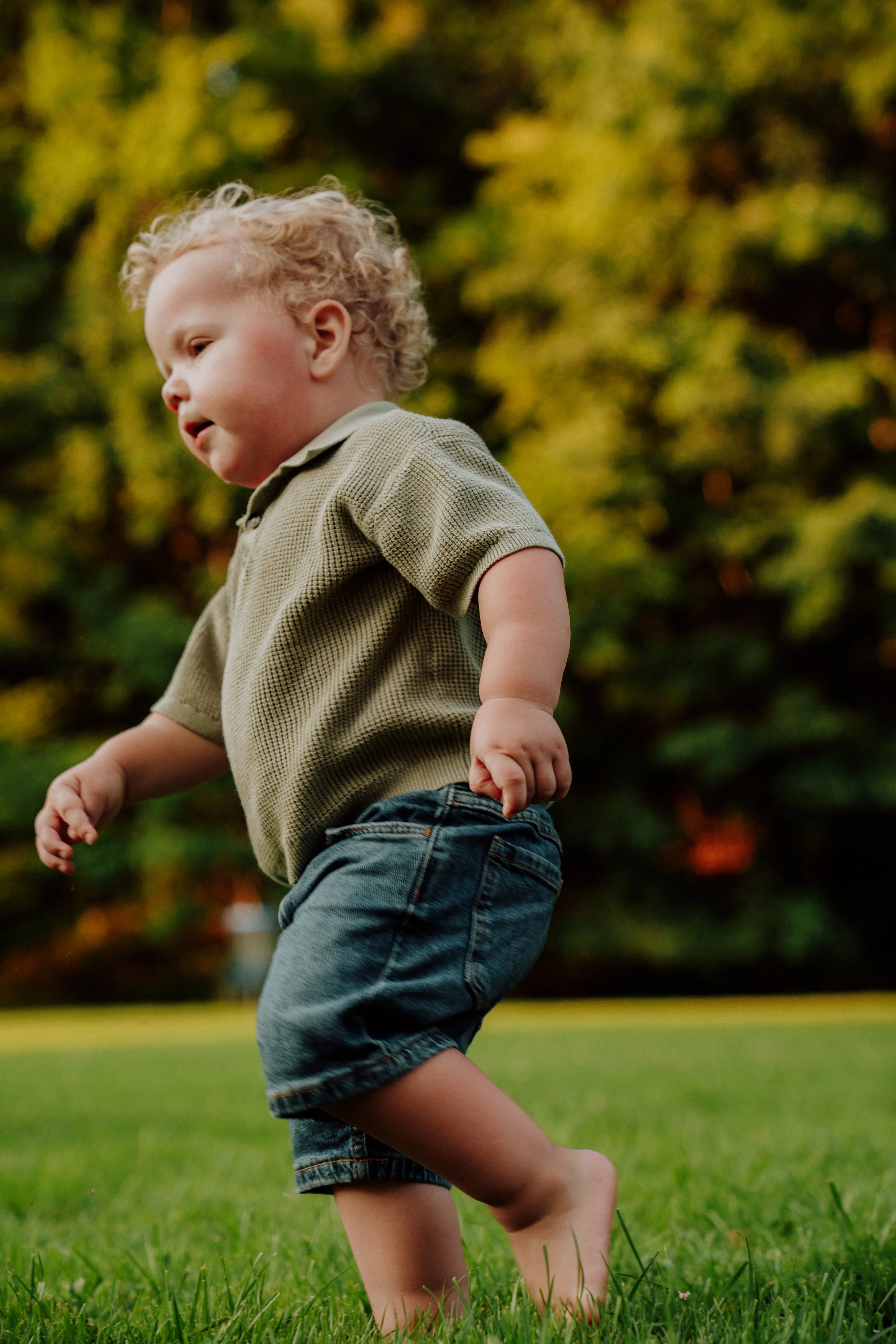 A young boy with curly blond hair wearing a green polo shirt and denim shorts, running on green grass with trees in the background.