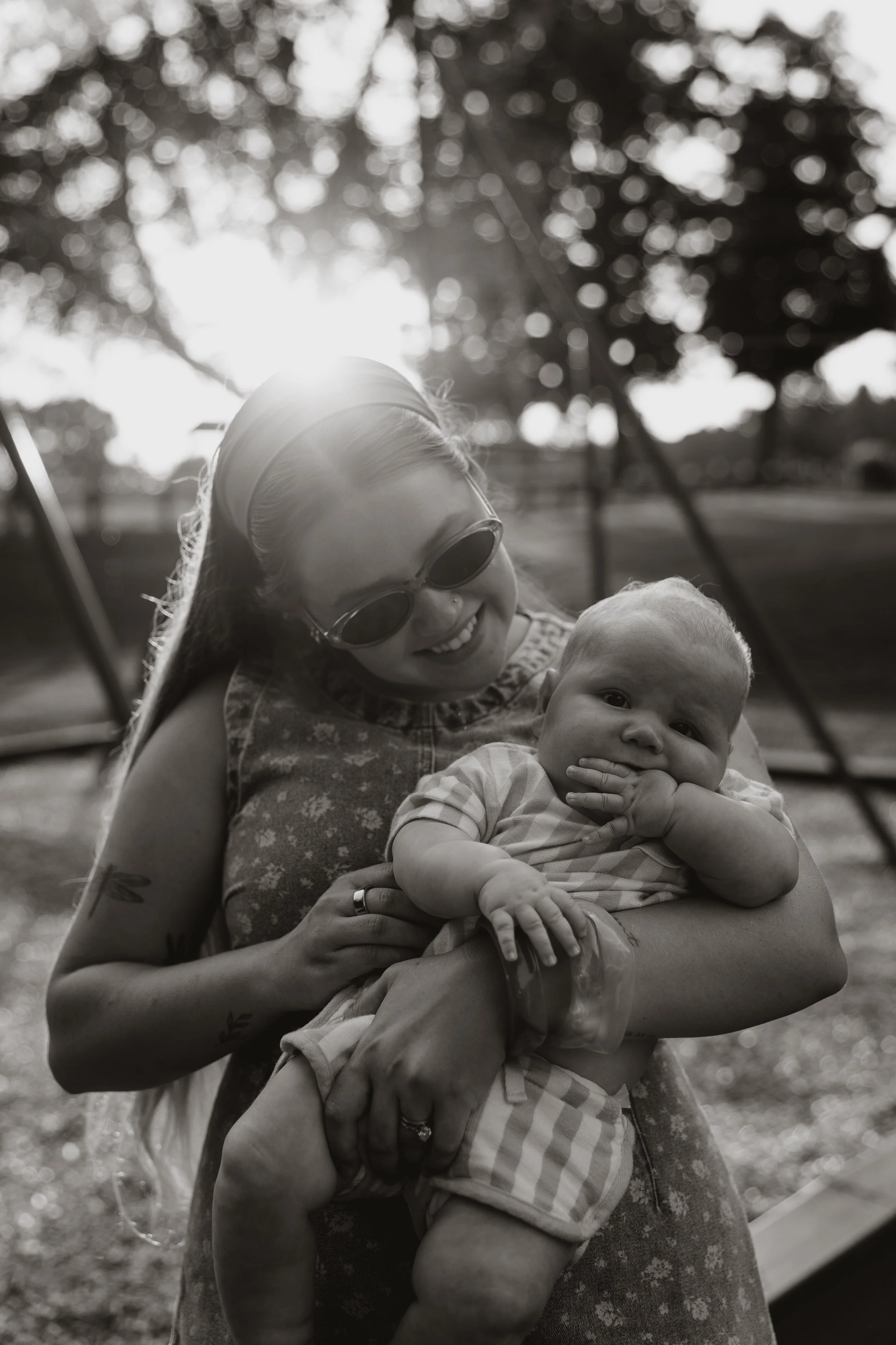 A woman with sunglasses holding a young baby outdoors, with trees and bright sunlight in the background.