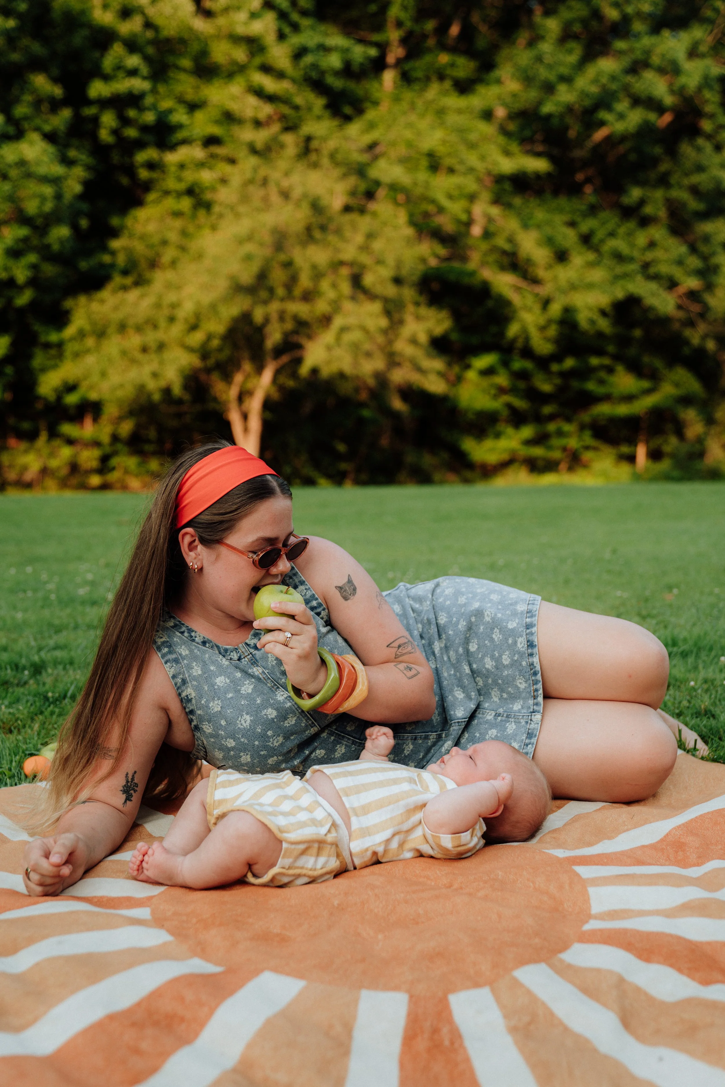 A woman with long hair, wearing sunglasses, a headband, and jewelry, lies on her side on a striped blanket on the grass in a park with trees in the background. She is holding a green apple and smiling. A baby with light hair, wearing a yellow striped