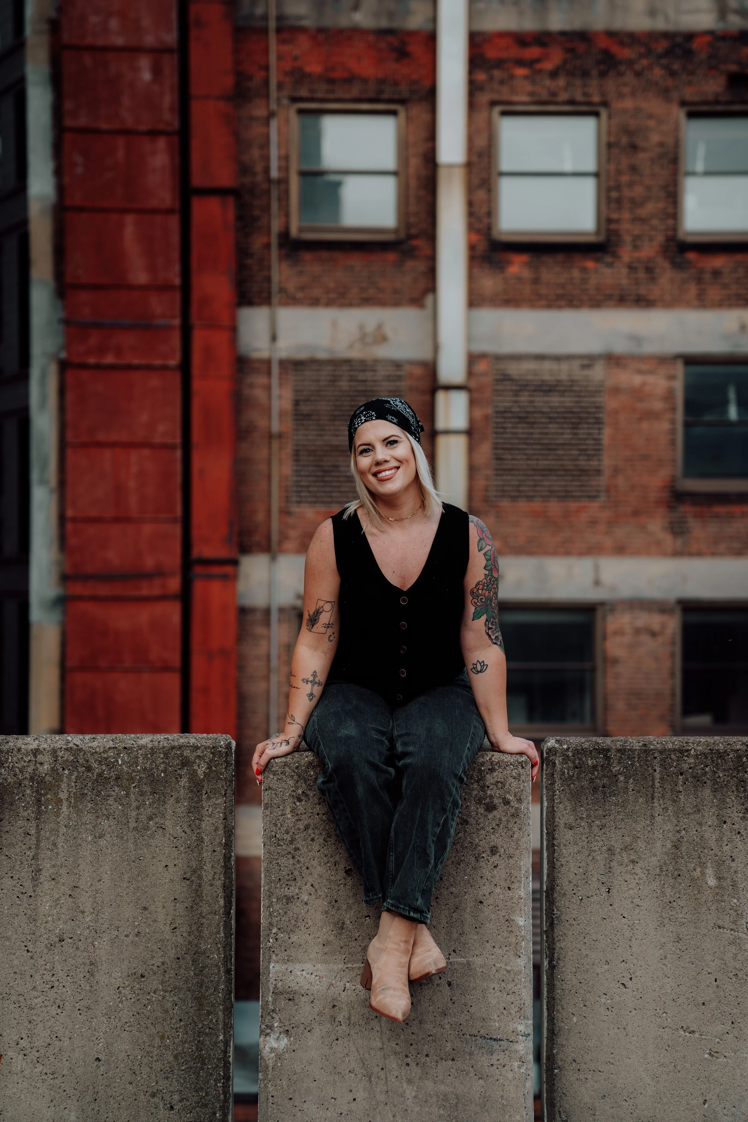 A woman sitting on a concrete block in front of a brick building, smiling, wearing a black sleeveless top, dark jeans, beige shoes, and a black bandana, with visible tattoos on her arms.