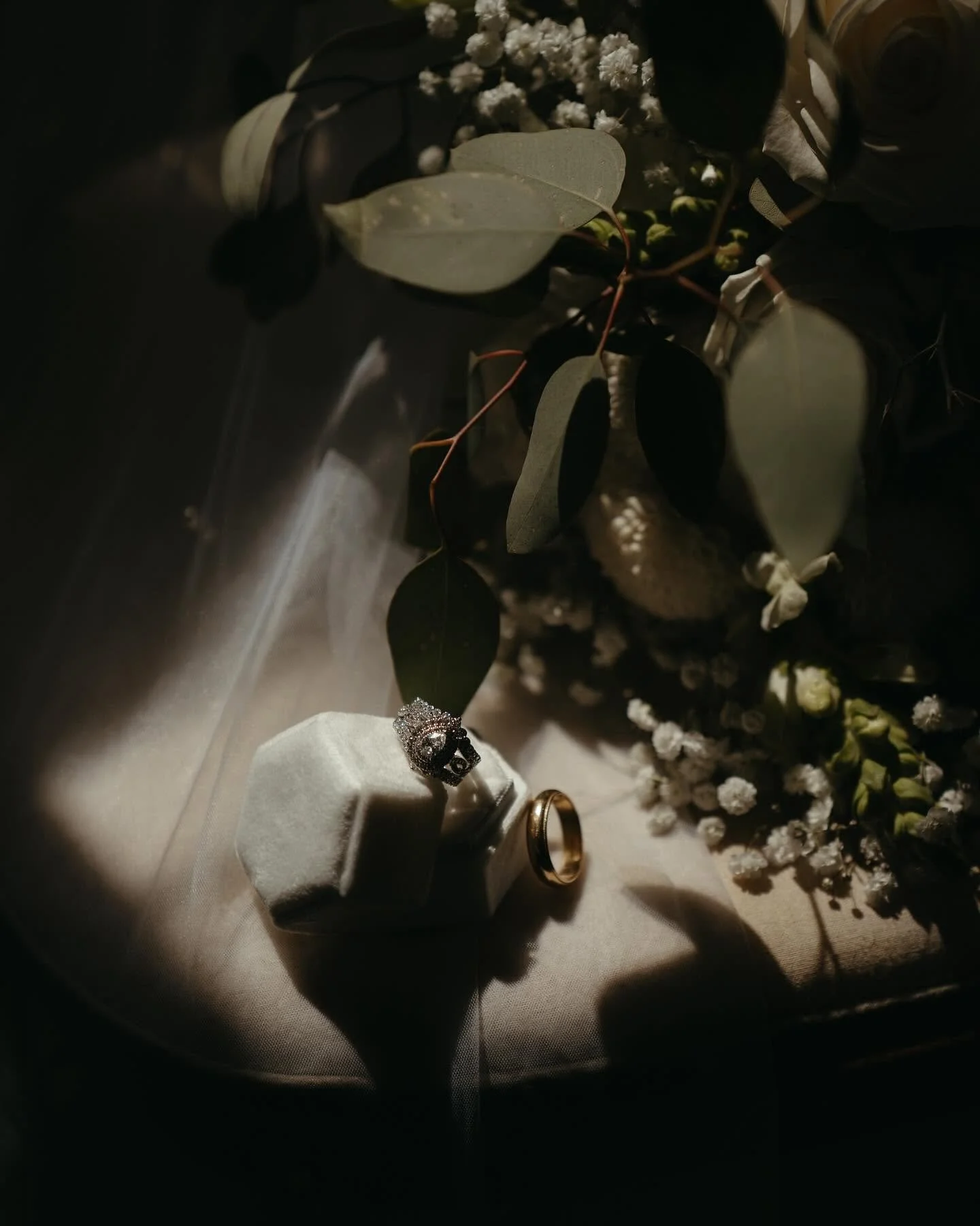 Diamond ring in a jewelry box, gold wedding band, and a floral arrangement of white baby's breath and green leaves in a dimly lit setting.