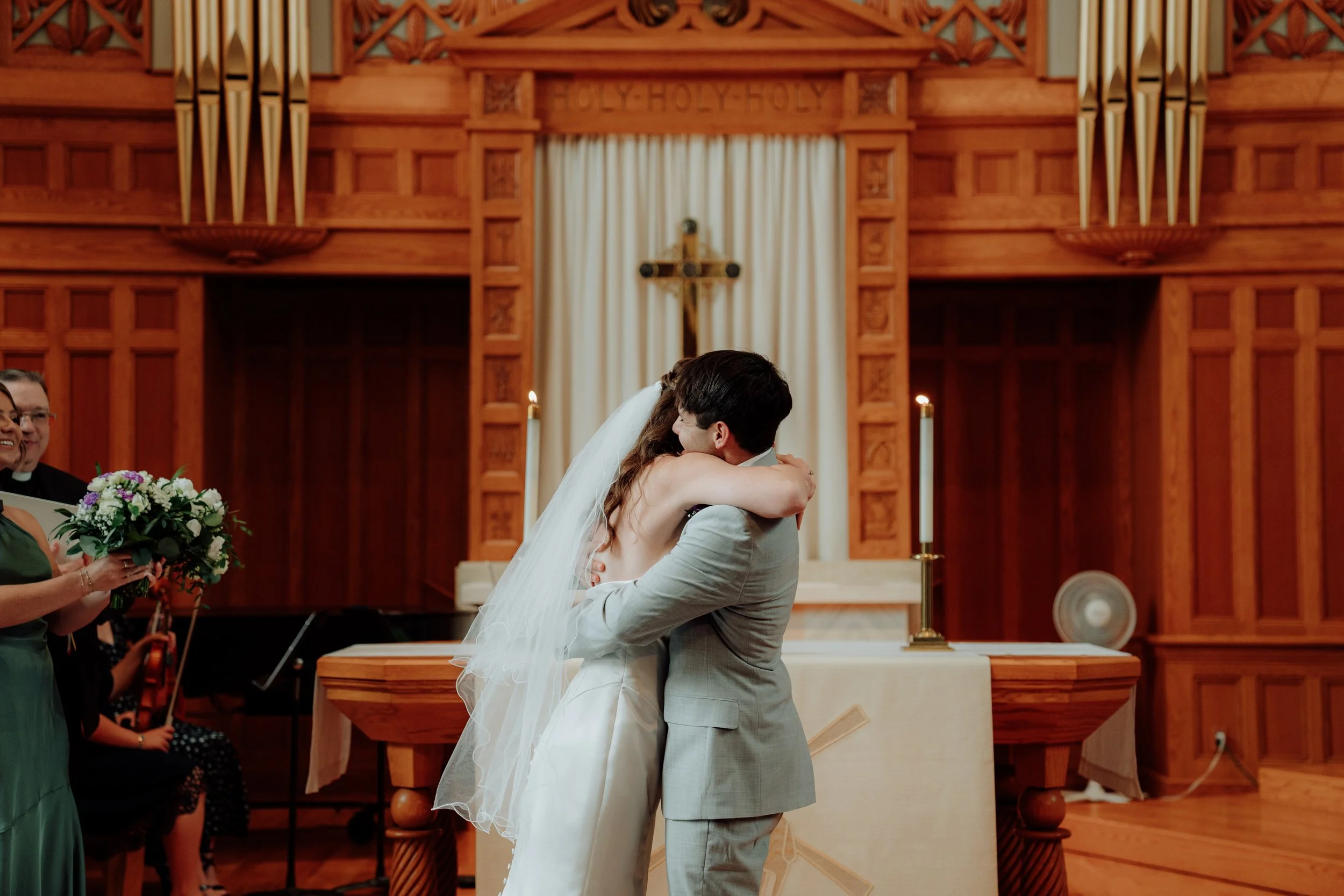 A bride and groom share a kiss at the altar in a church during their wedding ceremony. The church has wooden paneling and a crucifix behind them. A woman holds a bouquet of flowers to the left, and guests are visible in the background.