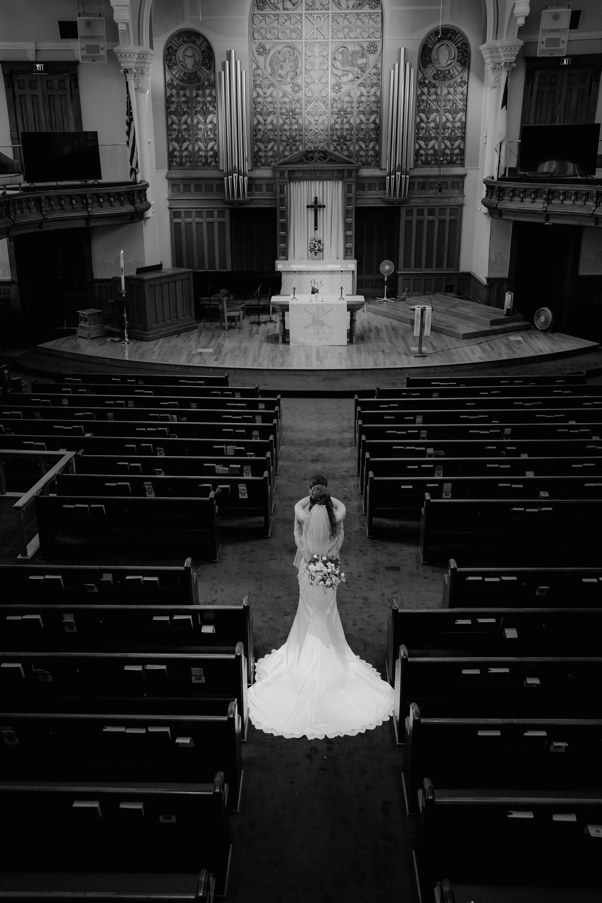A bride in a white wedding gown with a long train, holding a bouquet, is standing in a church aisle facing the altar, which has a cross, candles, and floral arrangement. The church interior features ornate woodwork, large pipe organ pipes, and two te