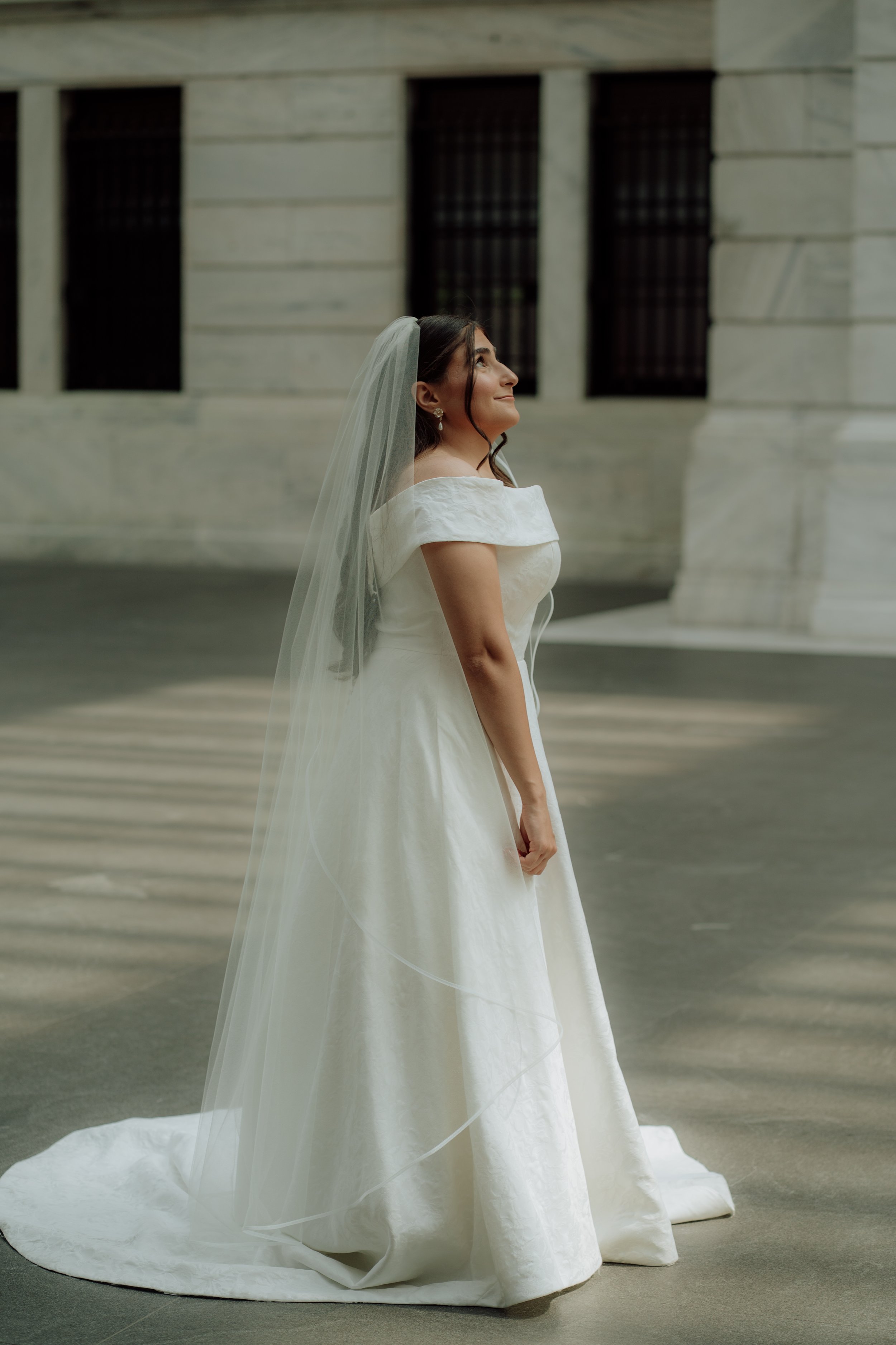 A bride in a white wedding dress with off-the-shoulder sleeves and a long train, standing outdoors in front of a stone building with large rectangular windows. She is smiling and looking up, wearing earrings and a veil.