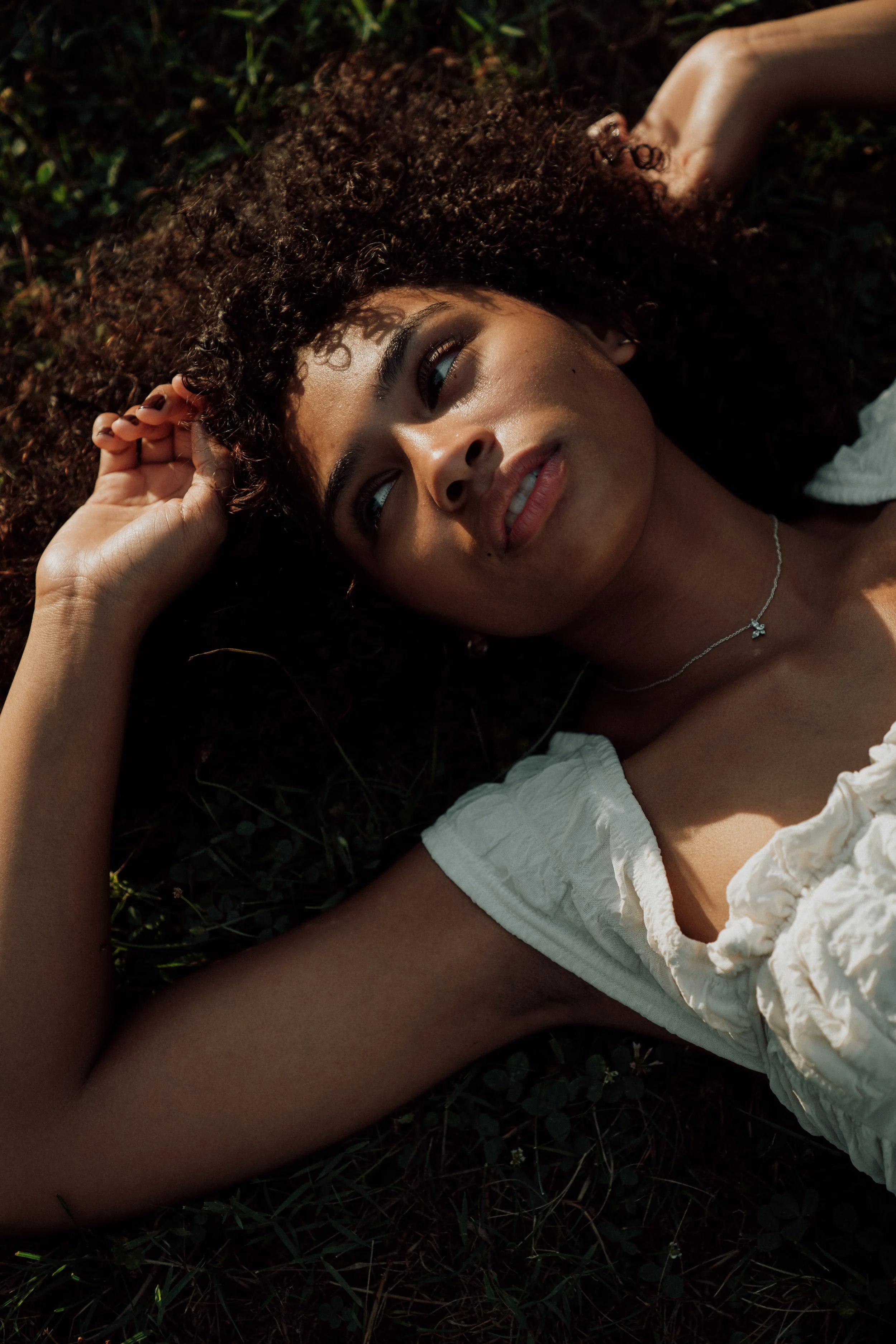 A woman with curly hair lying on the grass, wearing a white dress and a necklace.