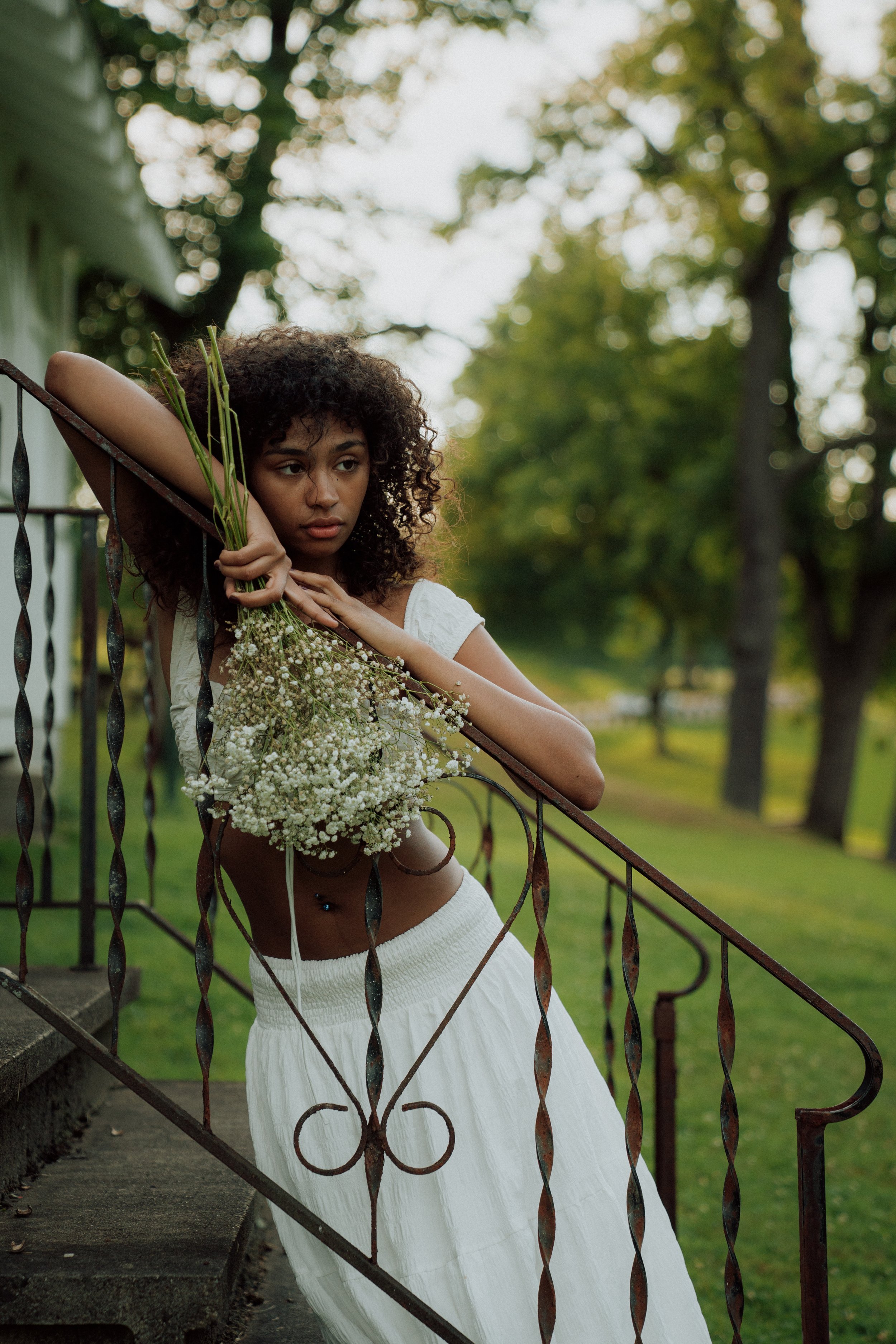 A young woman with curly hair leaning on a metal fence, holding a bouquet of white flowers, wearing a white skirt and a white top, outdoors with trees and greenery in the background.