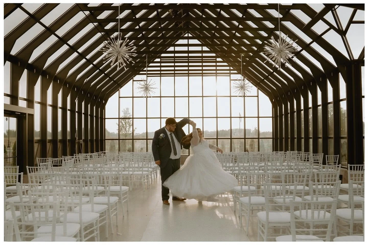 A bride and groom dancing inside a modern glass-walled wedding venue with empty white chairs arranged in rows.