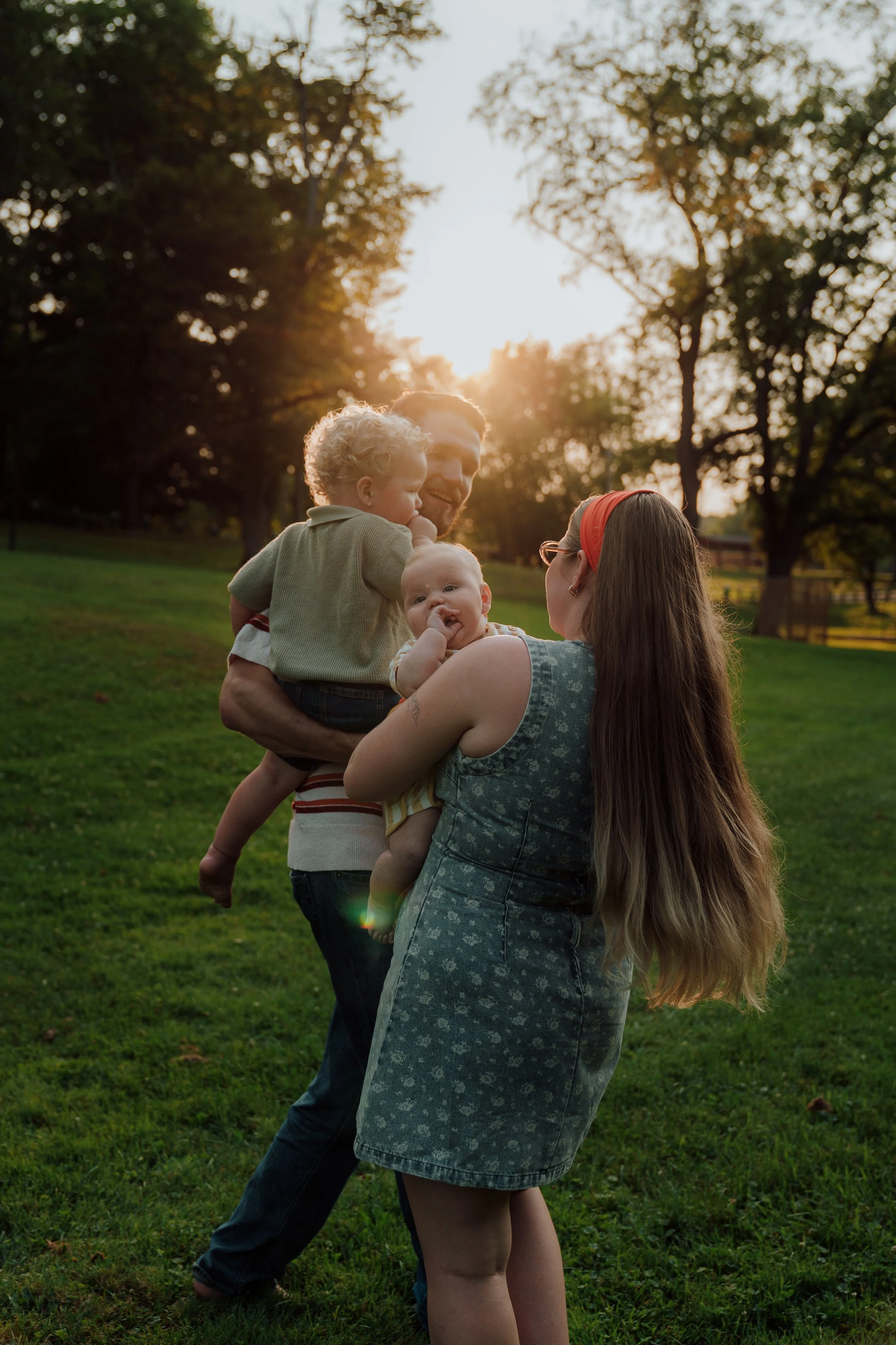 A family of four enjoying time together outdoors in a park during sunset, with trees and a wooden fence in the background.