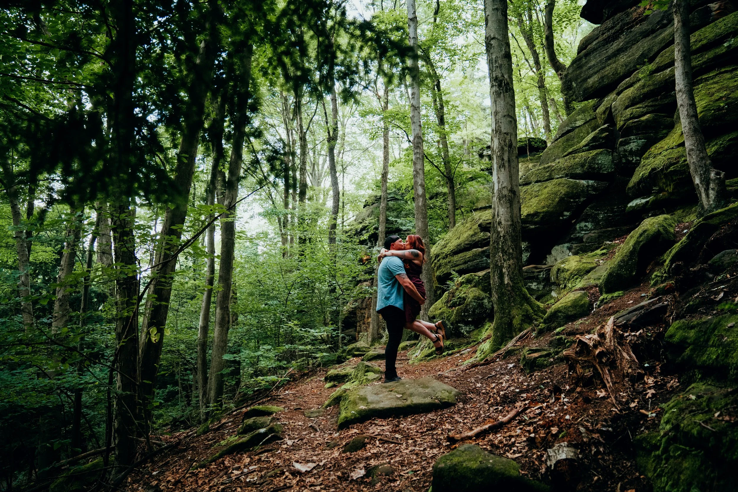 A couple sharing a kiss in a lush green forest. The woman is lifting her leg as the man holds her, near large moss-covered rocks and tall trees with bright green leaves.