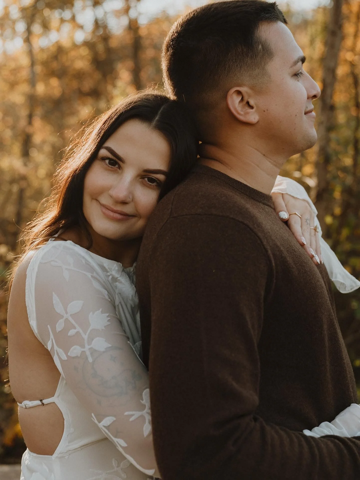 A couple embraces outdoors during autumn, with the woman smiling and resting her head on the man's shoulder, surrounded by trees with orange and yellow leaves.